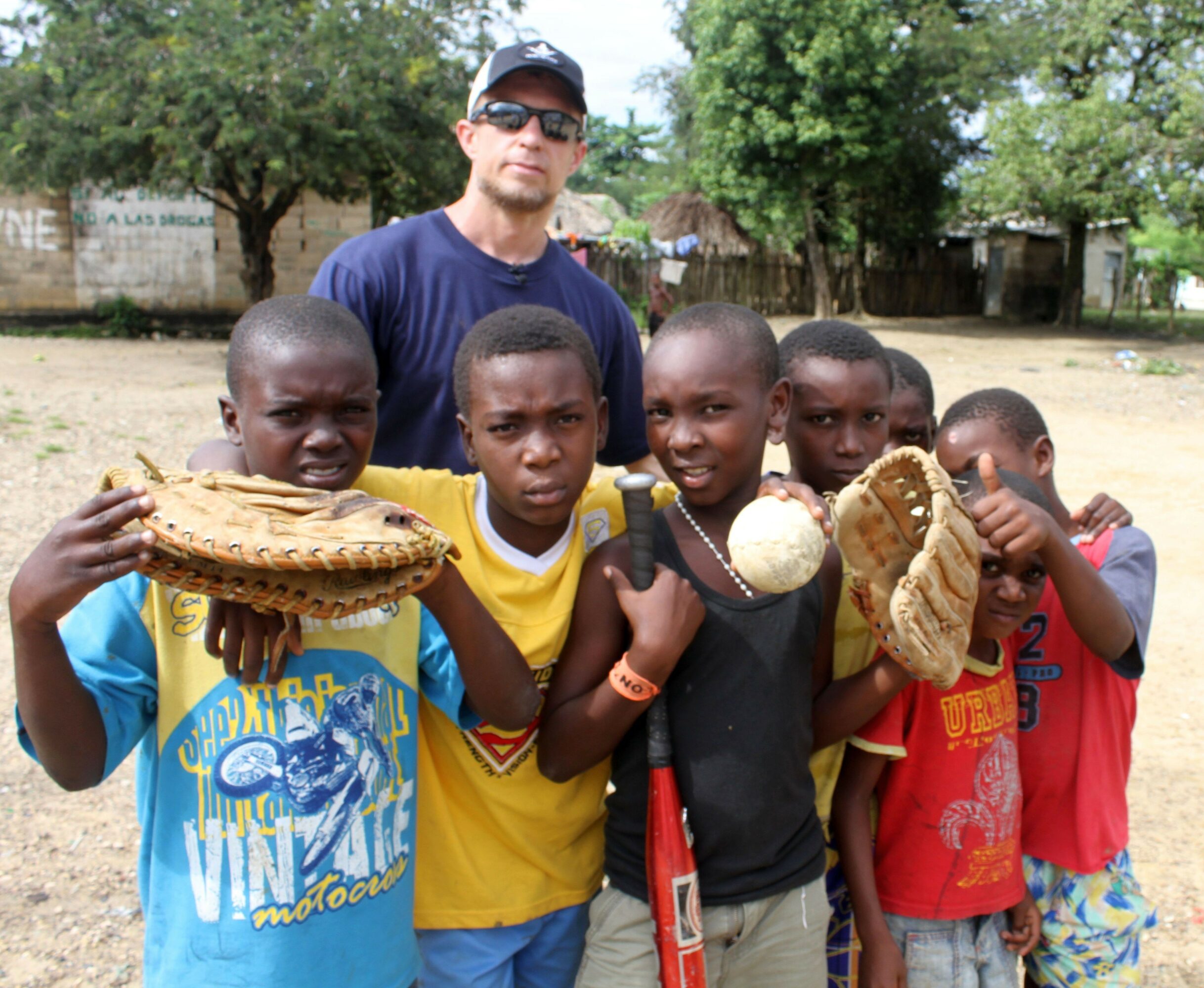 Group of five children holding baseball gloves and a ball, with a man standing behind them outdoors.