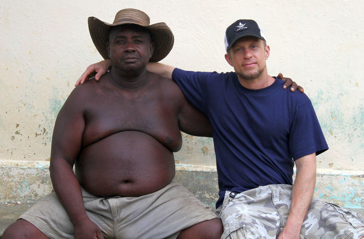 Two men sit side by side, one shirtless wearing a hat, the other in a navy shirt and cap, against a plain wall.