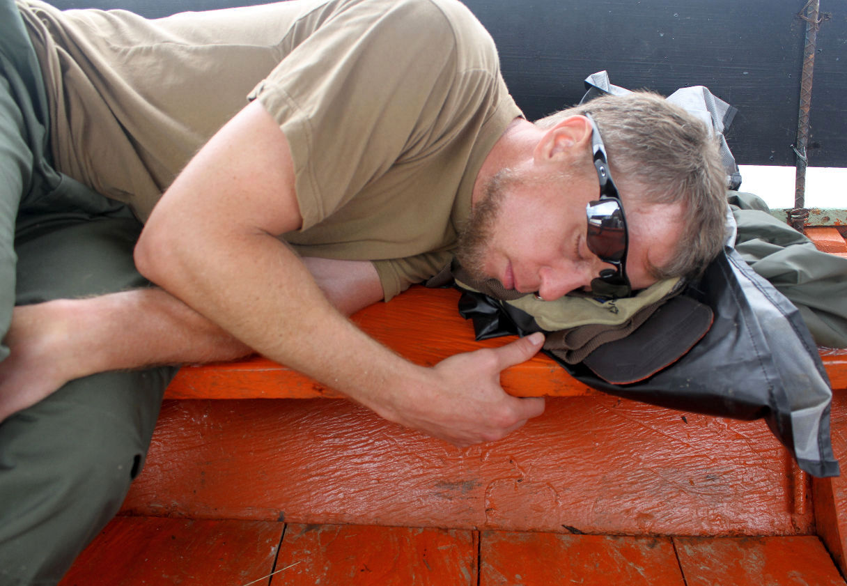 Man with sunglasses lying on a wooden surface, resting his head on a backpack, with a beige shirt and green pants.