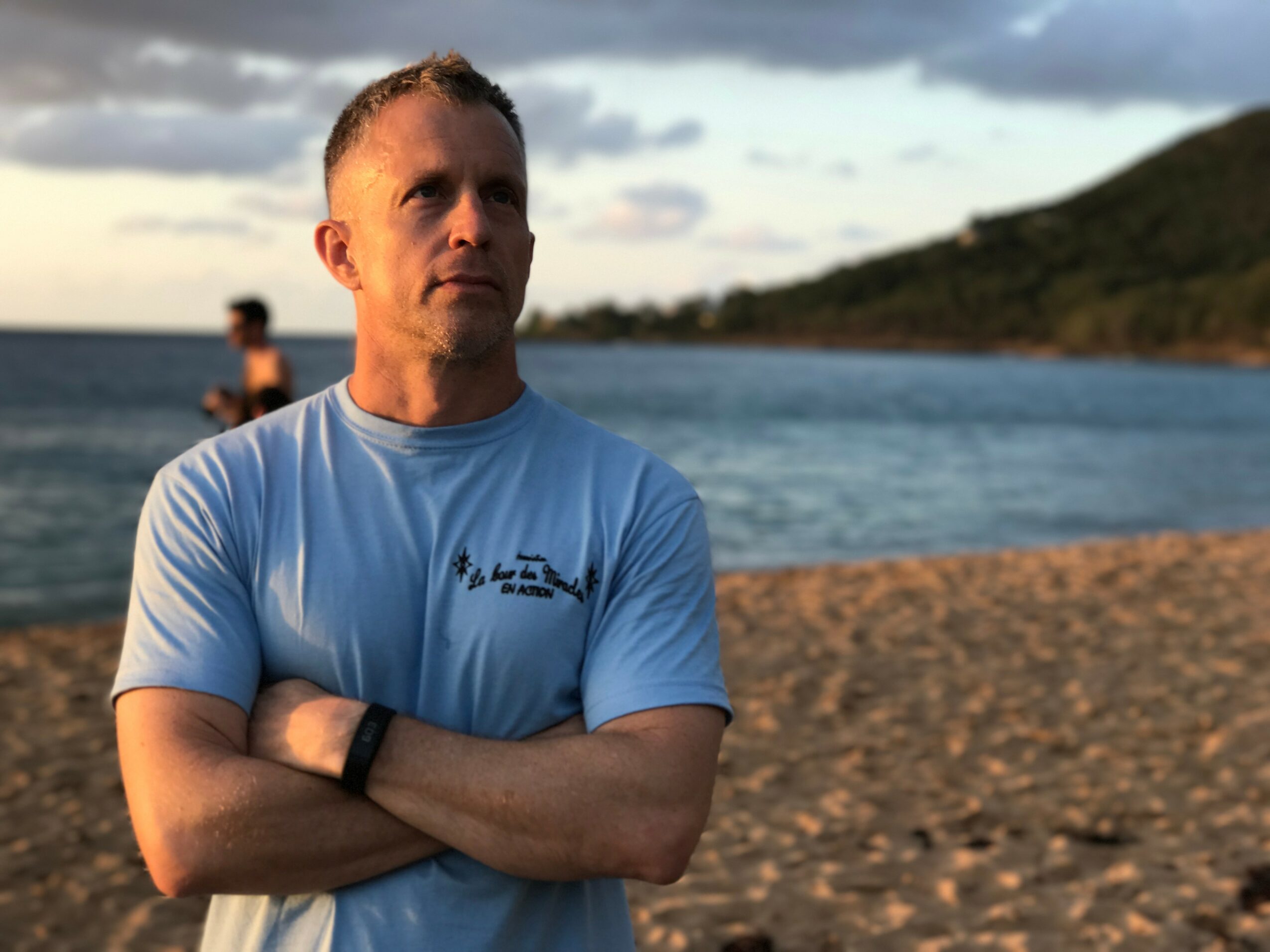 Man with arms crossed standing on a beach with water and hills in background, during sunset.