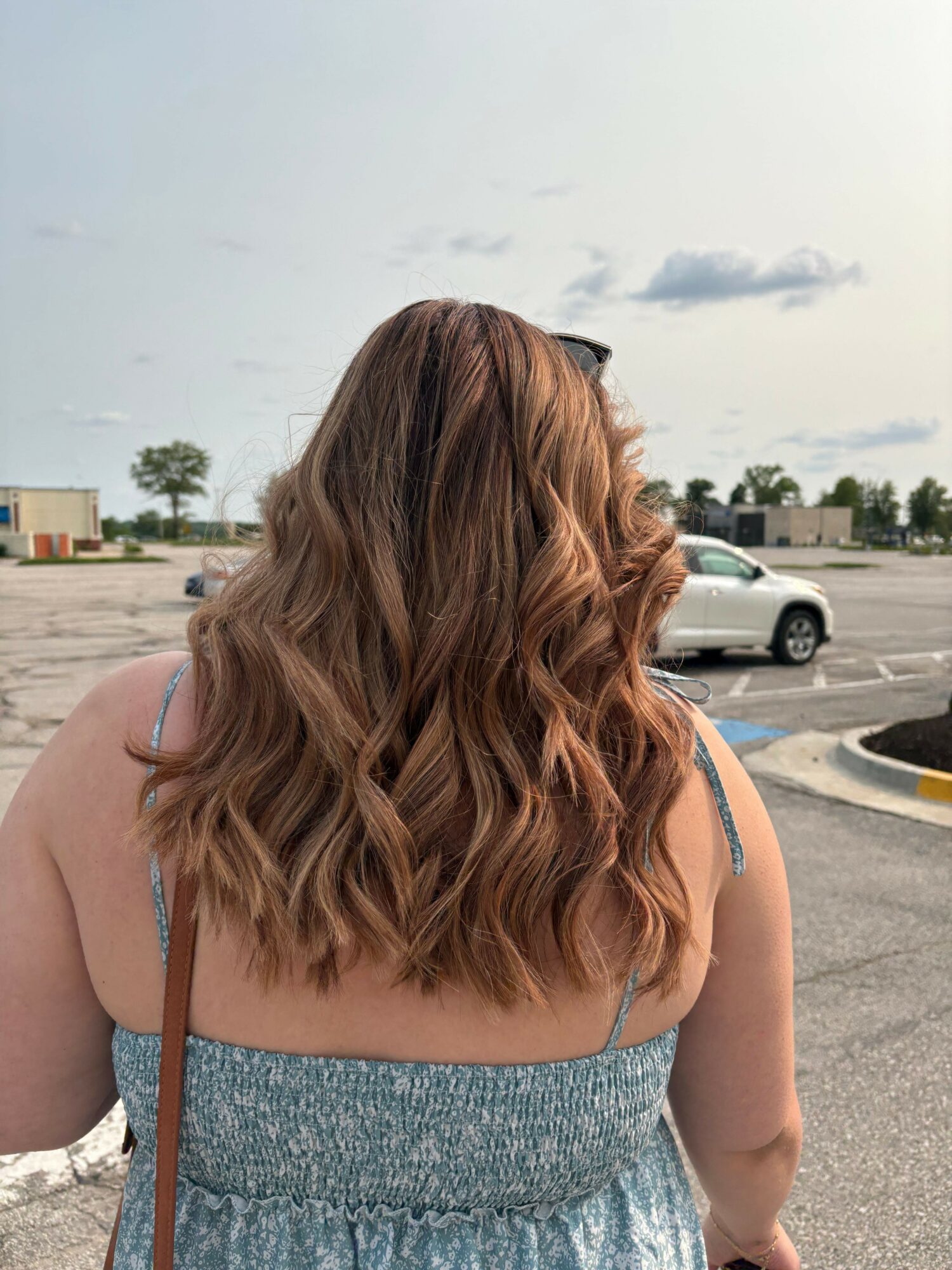 Person with shoulder-length wavy hair wearing a sleeveless top, standing outdoors near parked cars.