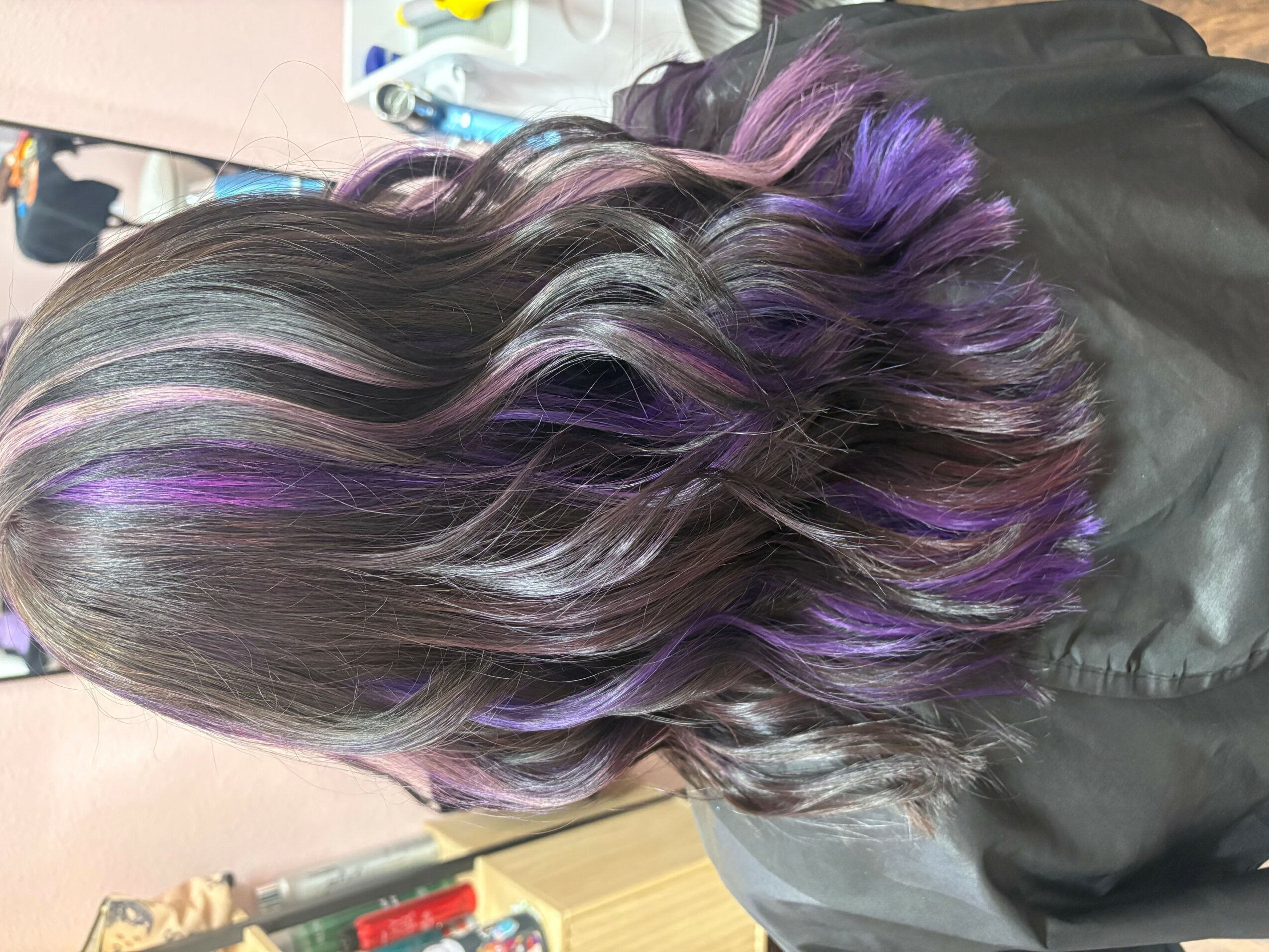 Back view of a person's wavy hair with purple highlights, resting on a black surface.