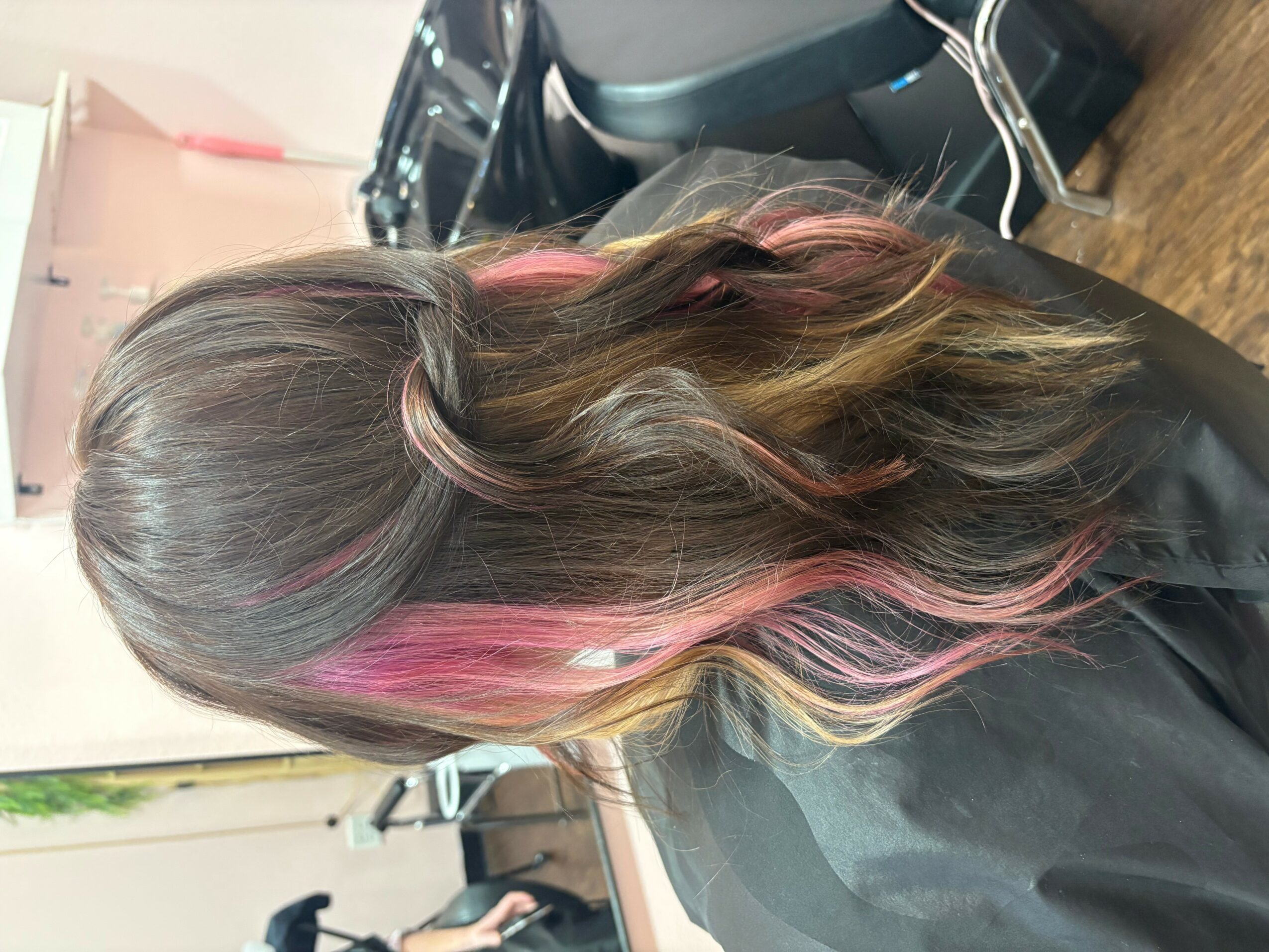 Side view of a person with long, wavy hair featuring pink highlights, sitting in a salon chair.