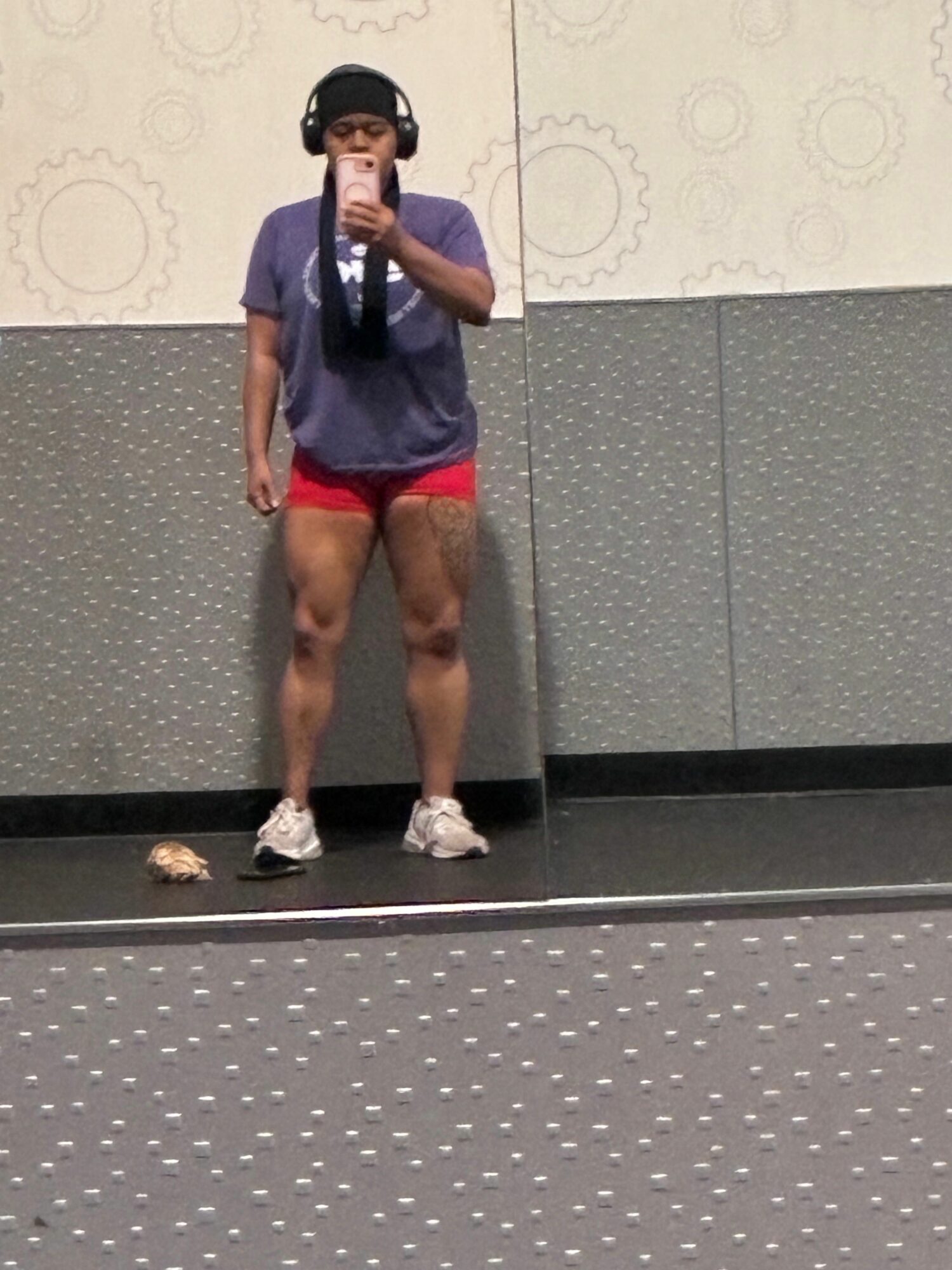 Person taking a mirror selfie in a gym, wearing a purple shirt, red shorts, and sneakers, with a phone in hand.
