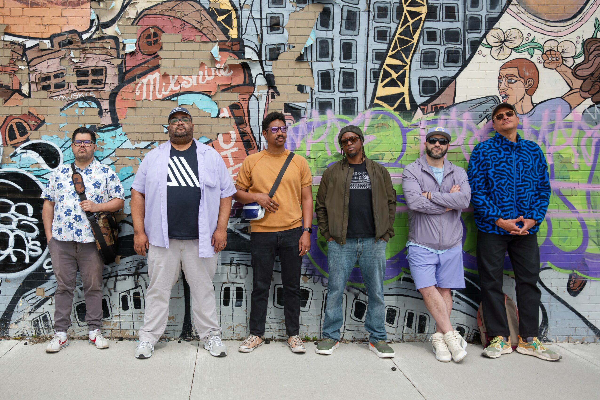 Six people stand in front of a colorful graffiti wall, posing for a photo outdoors.
