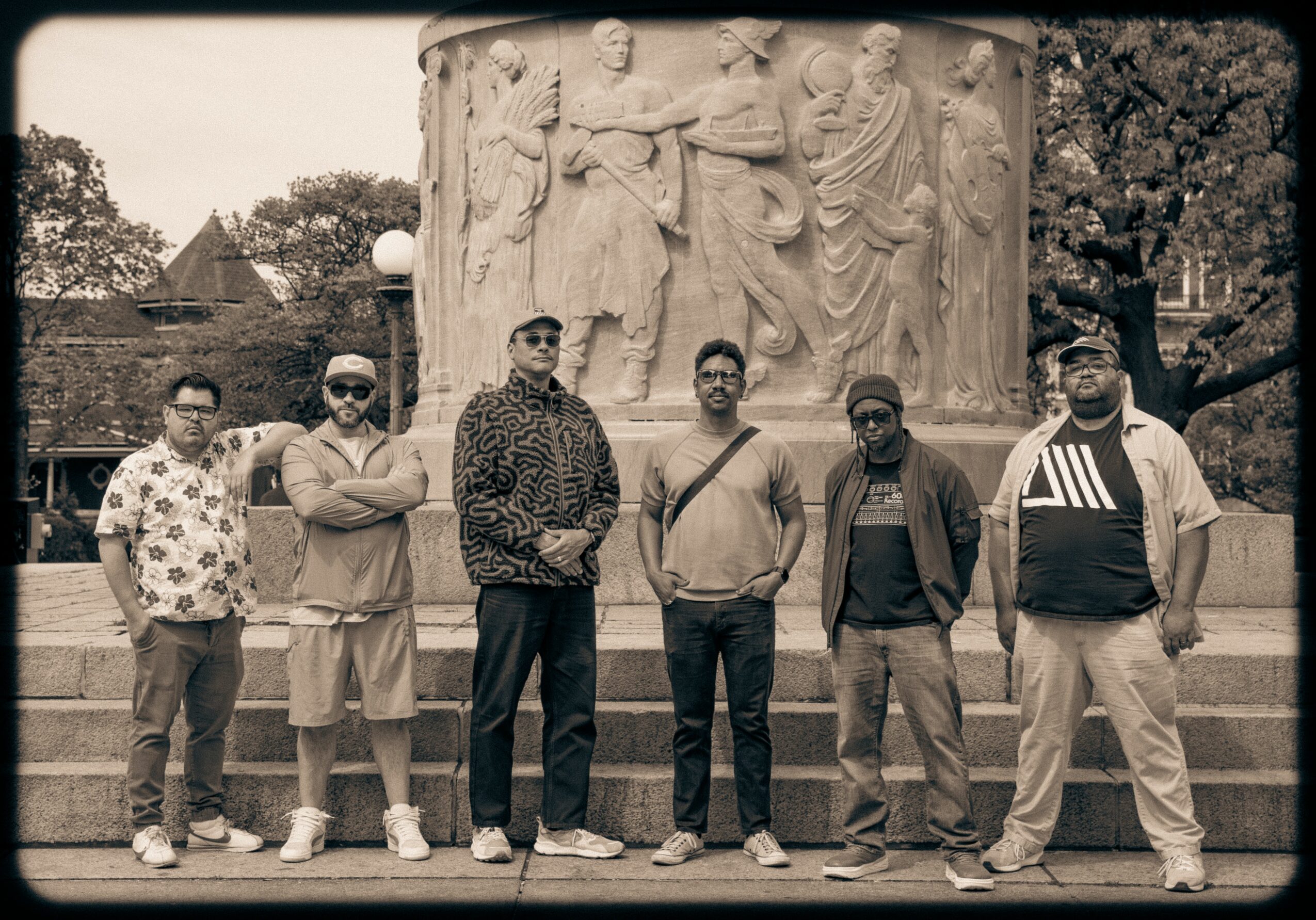 Six people stand in front of a large stone sculpture with carved figures, outdoors with trees and buildings in background.
