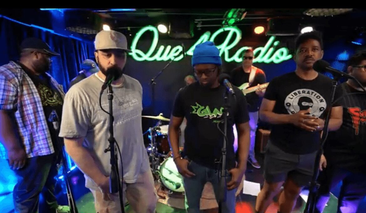 Group of five people on stage at Que Radio, with a drum set and neon sign in background.