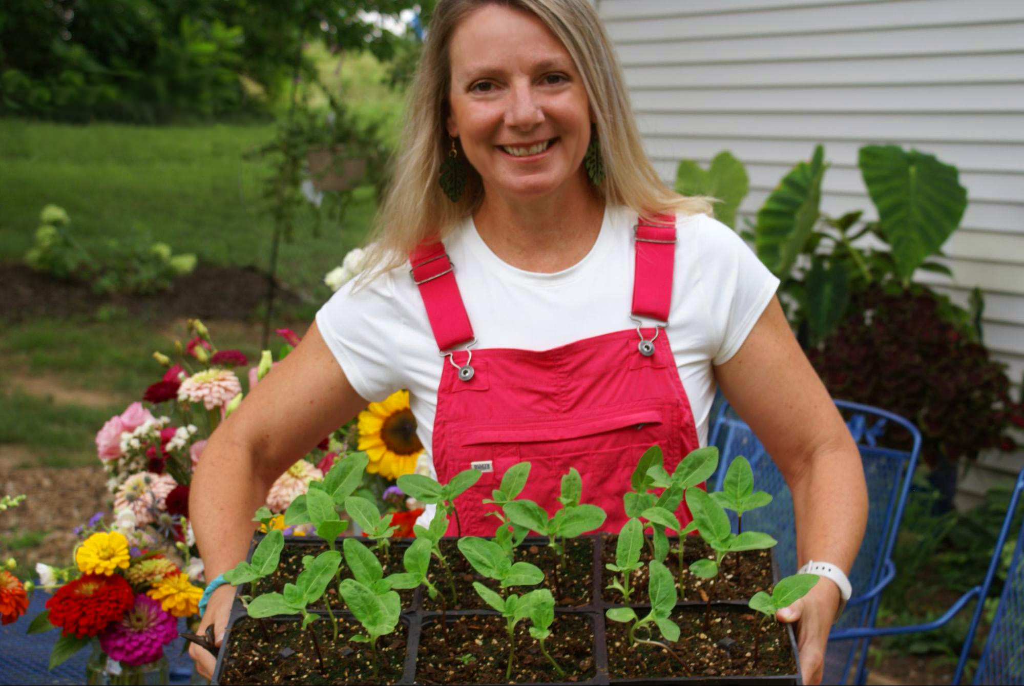 Woman with long blonde hair smiling outdoors, holding potted plants, garden with flowers and greenery in background.