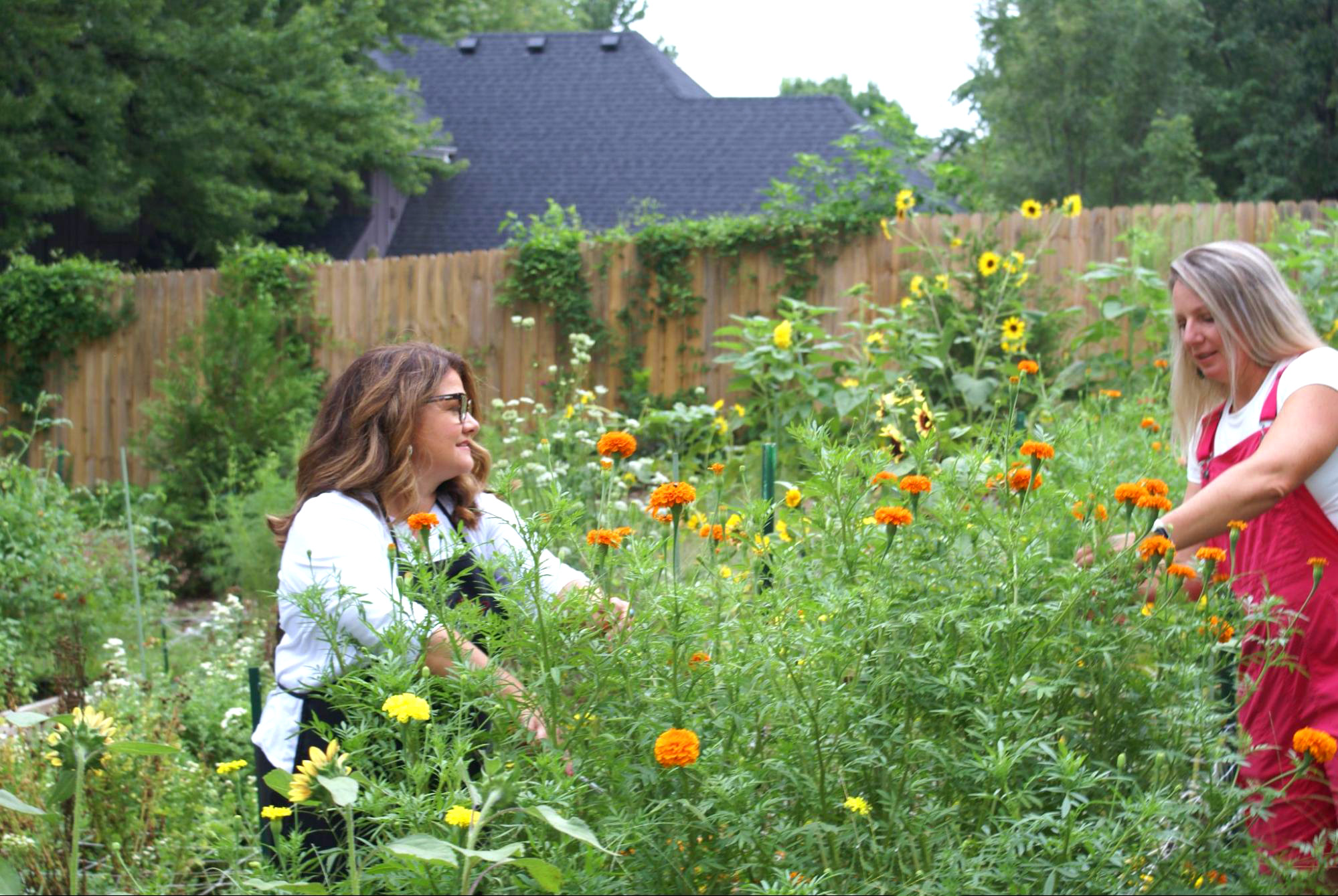 Two women in a garden with colorful flowers, one with glasses and brown hair, the other with blonde hair, smiling.