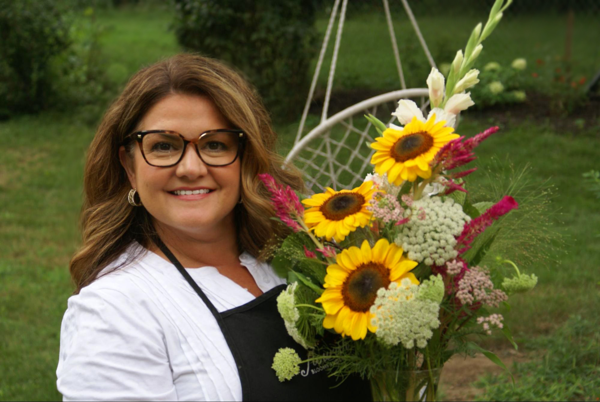 Woman with glasses holding a colorful flower arrangement outdoors, smiling, with green trees and grass in background.