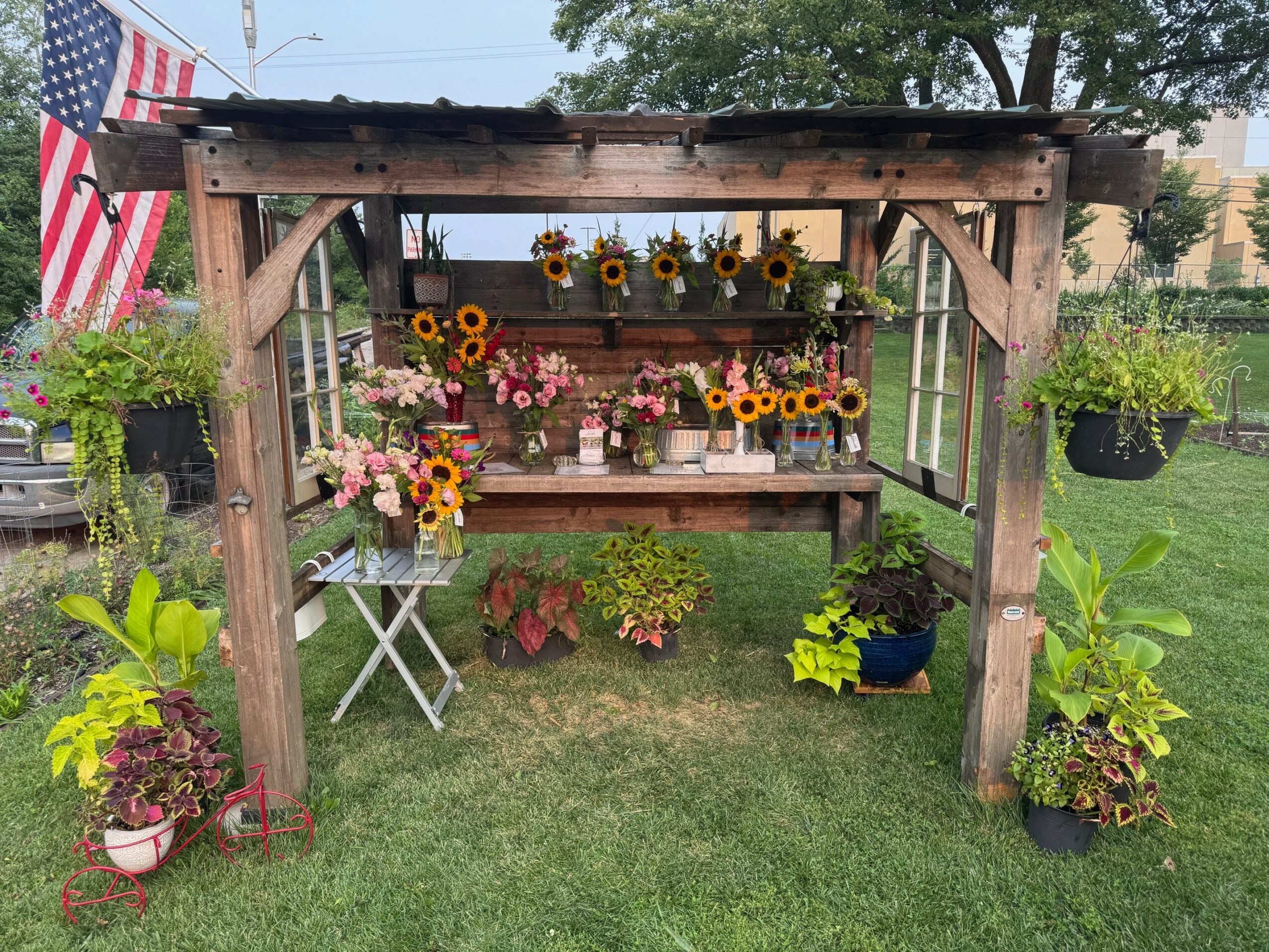 Wooden garden structure with hanging and potted flowers, sunflowers, and greenery, on a grassy lawn.
