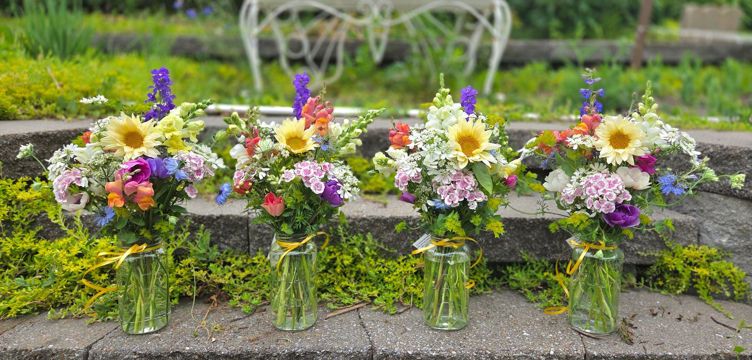 Five glass jars with colorful flower arrangements on a stone surface, with a garden and white metal furniture in the background.