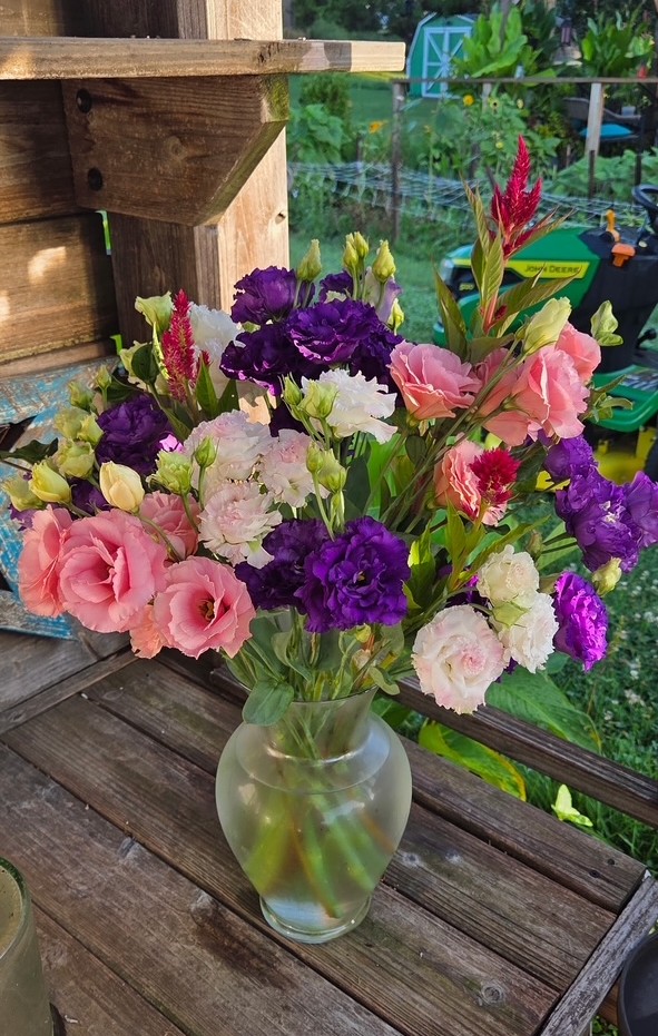 Vase with pink, purple, and white flowers on wooden surface outdoors, greenery and a fence in background.