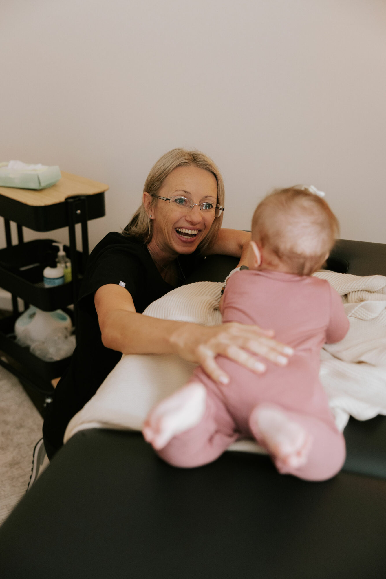 Woman smiling and reaching out to a baby on a hospital bed, with medical supplies nearby.