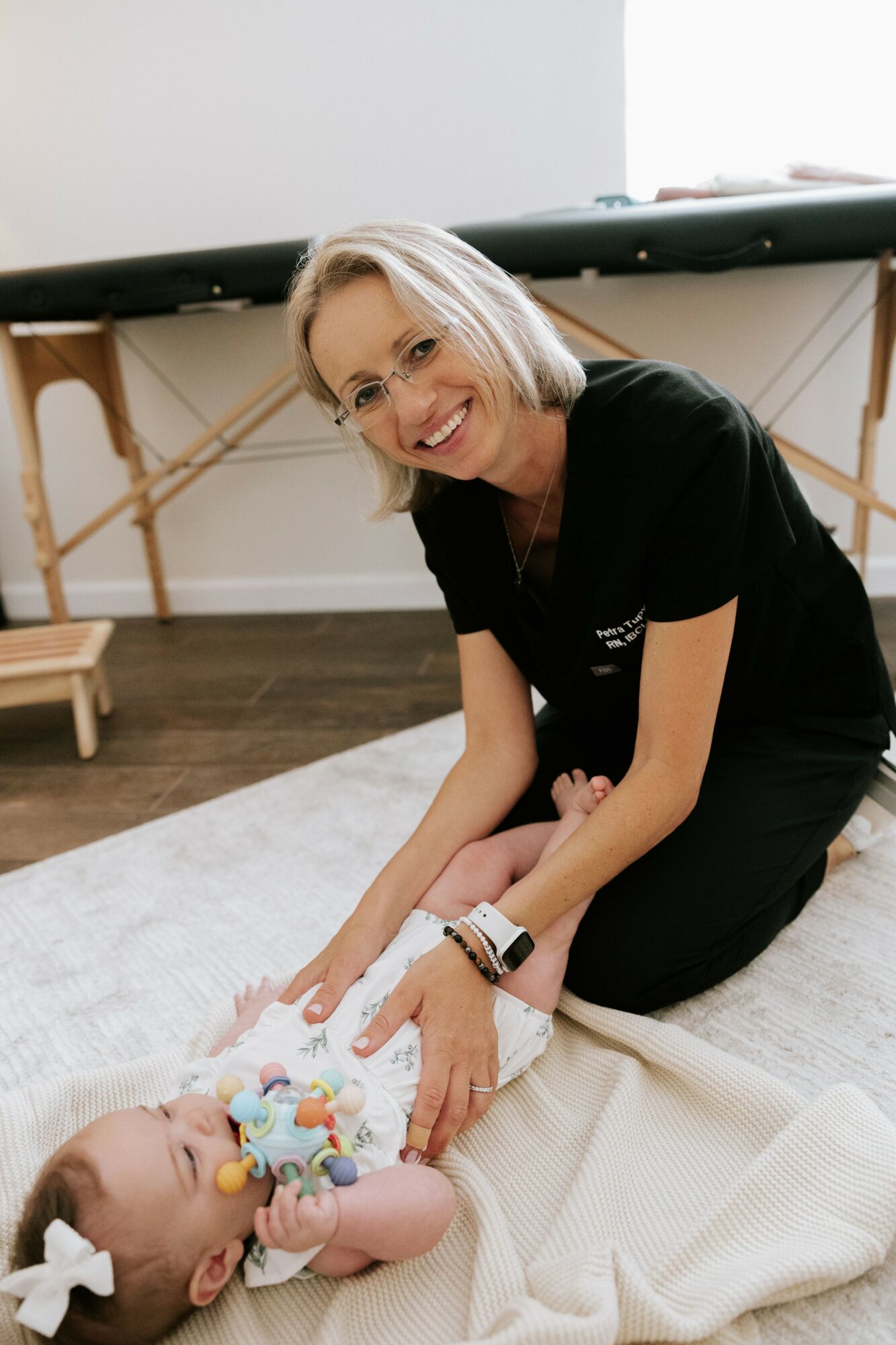 Woman smiling and playing with a baby lying on a blanket indoors, with a table in the background.