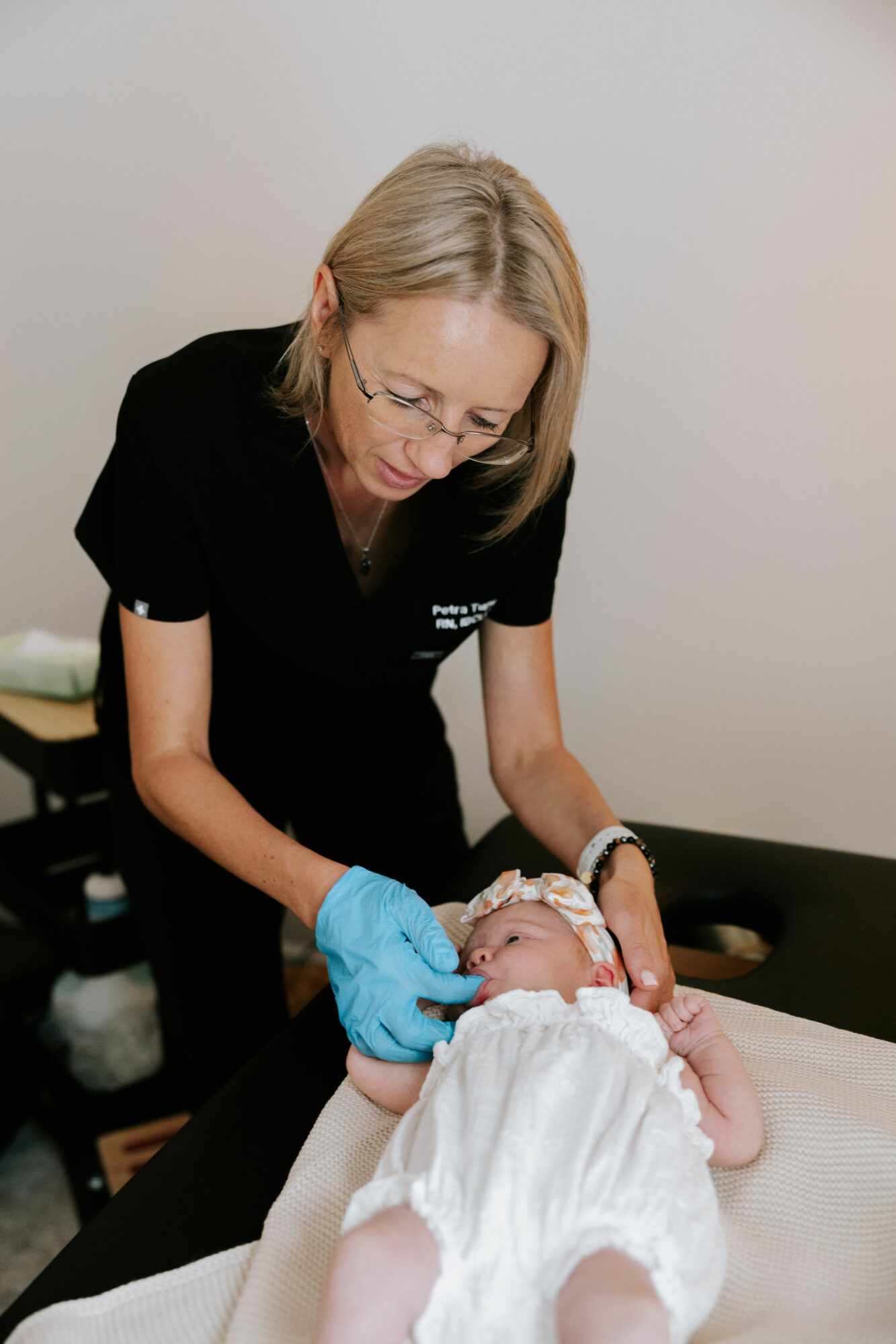 A woman wearing glasses and a black uniform cares for a baby lying on a table, holding the baby's hand with blue gloves.