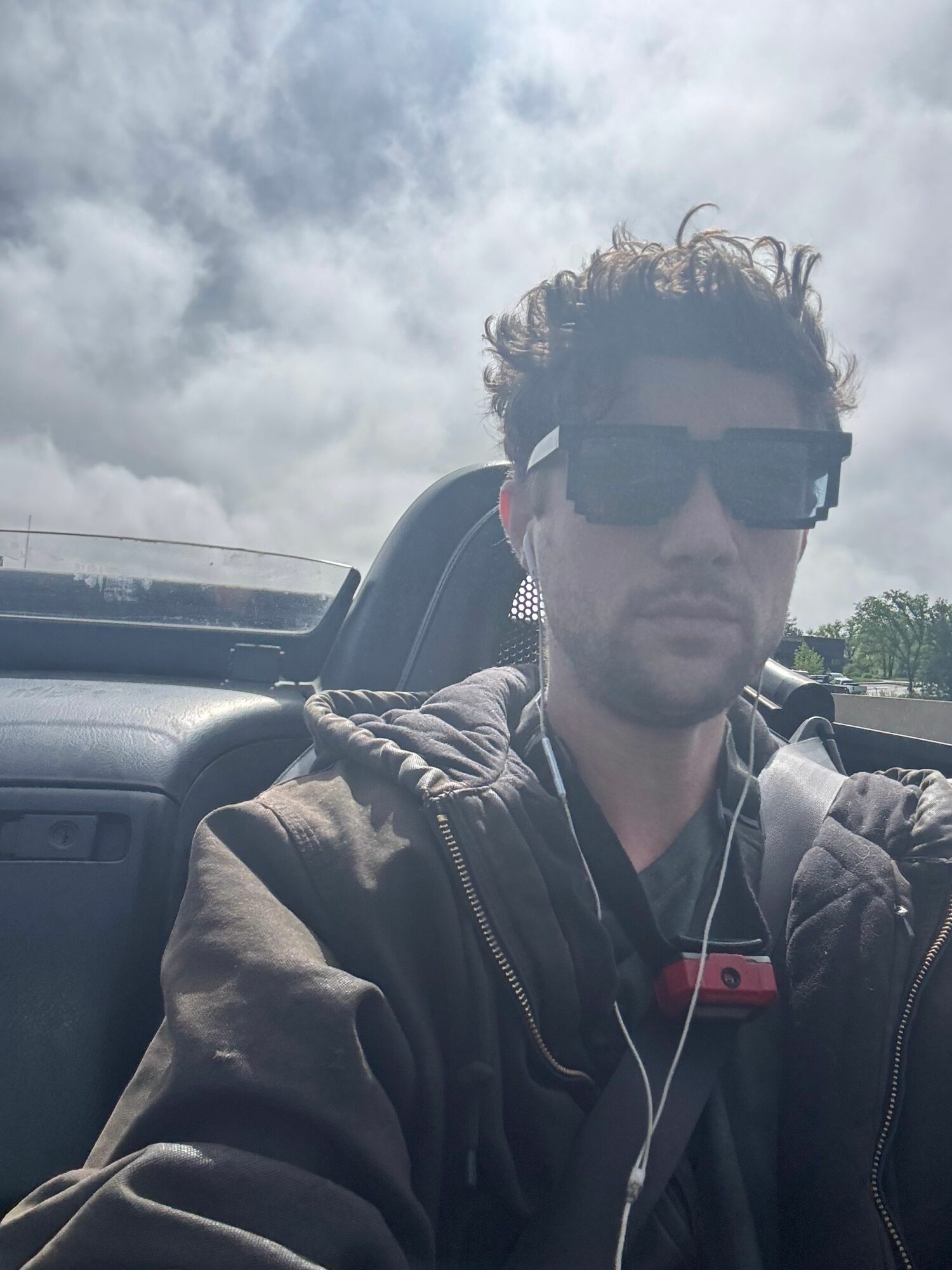 Young man with curly hair wearing sunglasses, headphones, and a jacket outdoors on cloudy day.
