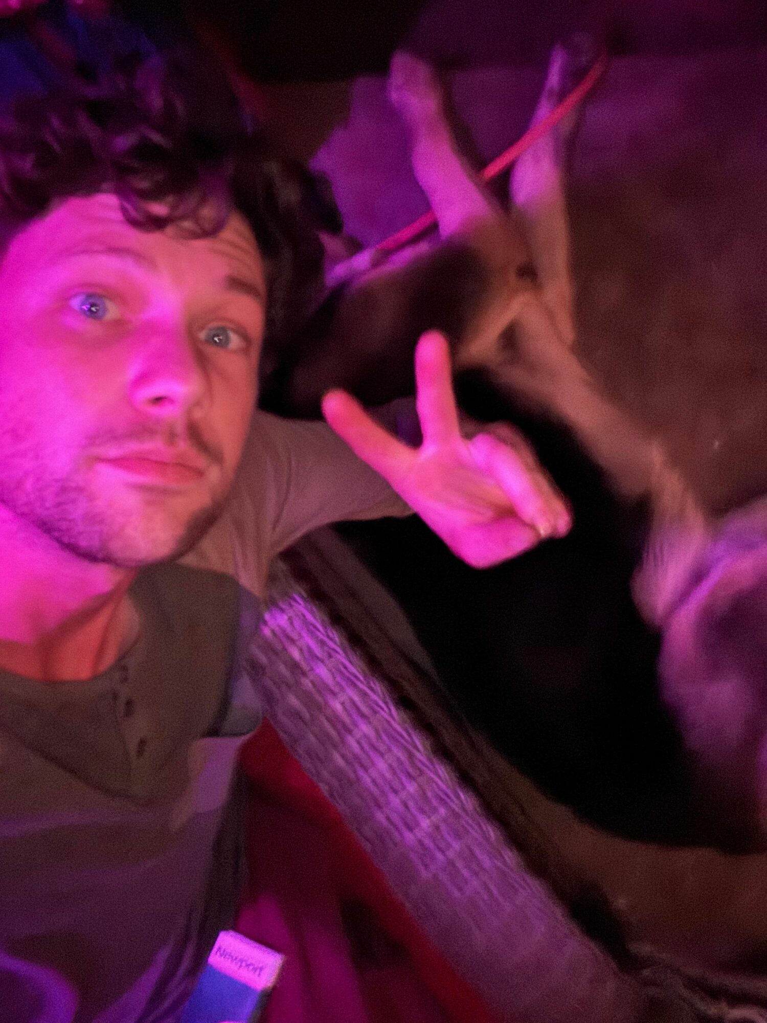 Person with curly hair lying down with a dog, both illuminated by pinkish light, in a cozy setting.