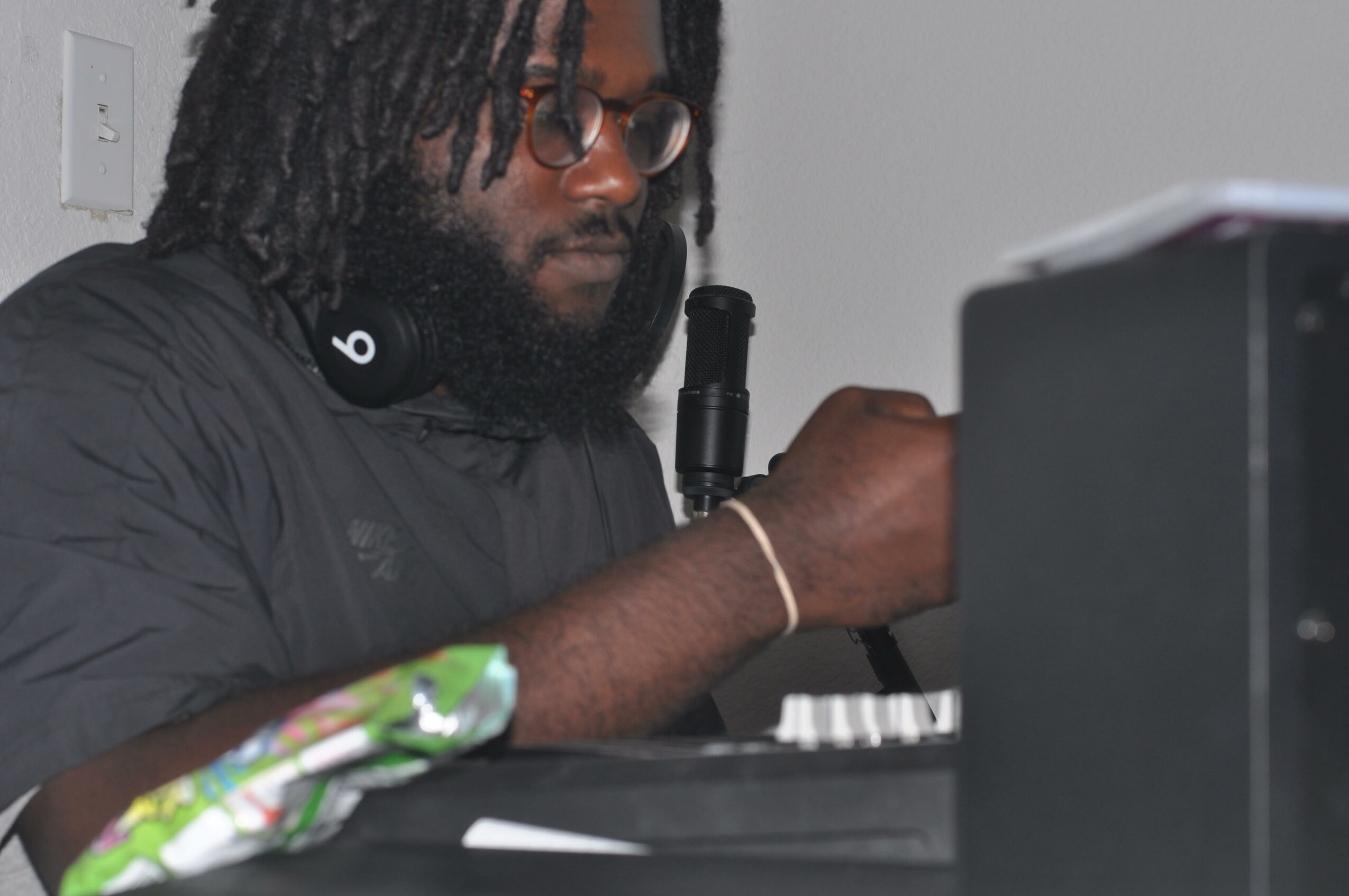 Person with dreadlocks and glasses working at a desk with electronic equipment, wearing headphones around neck.