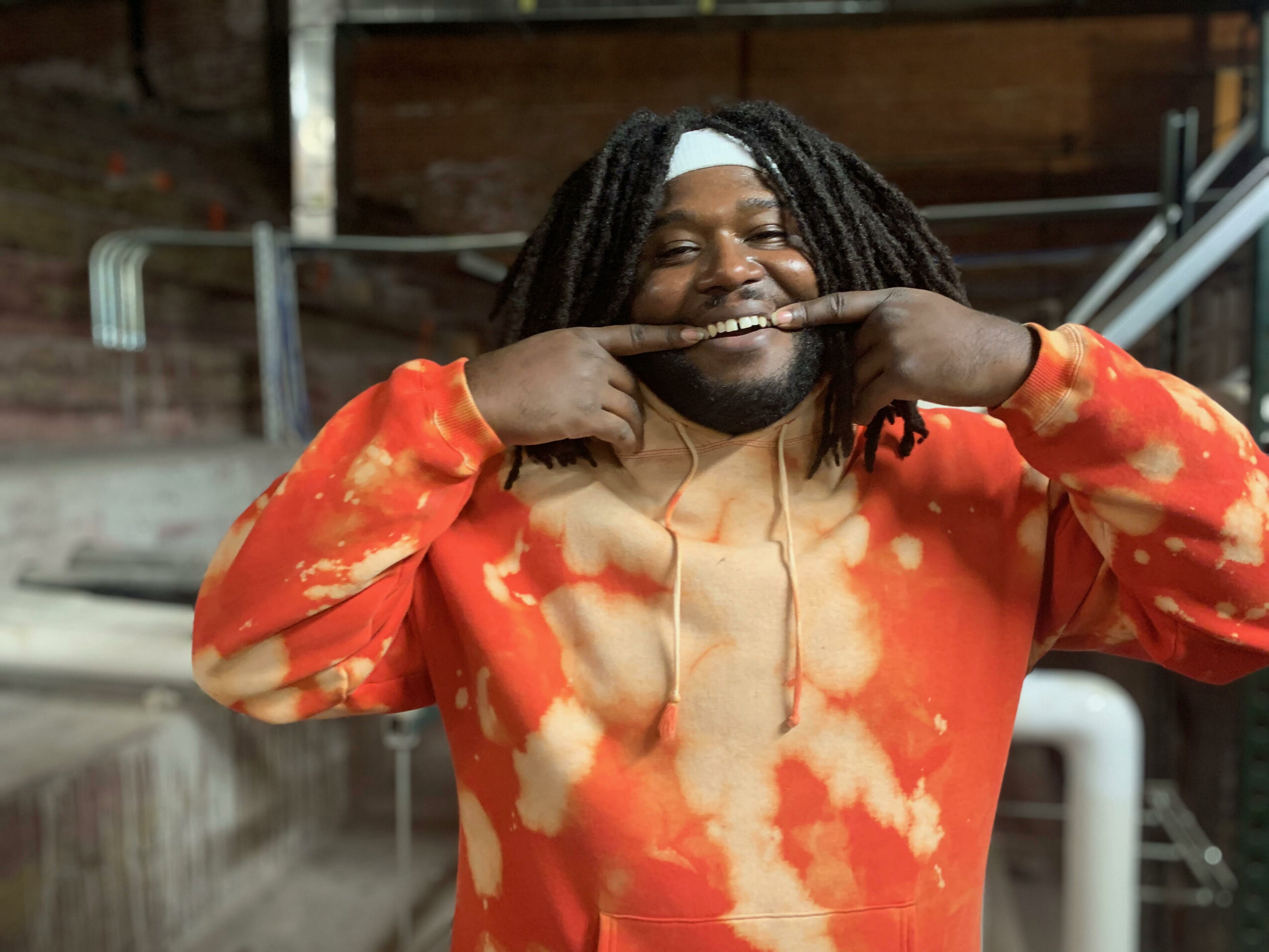 Person with dreadlocks smiling, wearing an orange and beige tie-dye hoodie, making a playful gesture with fingers on face.