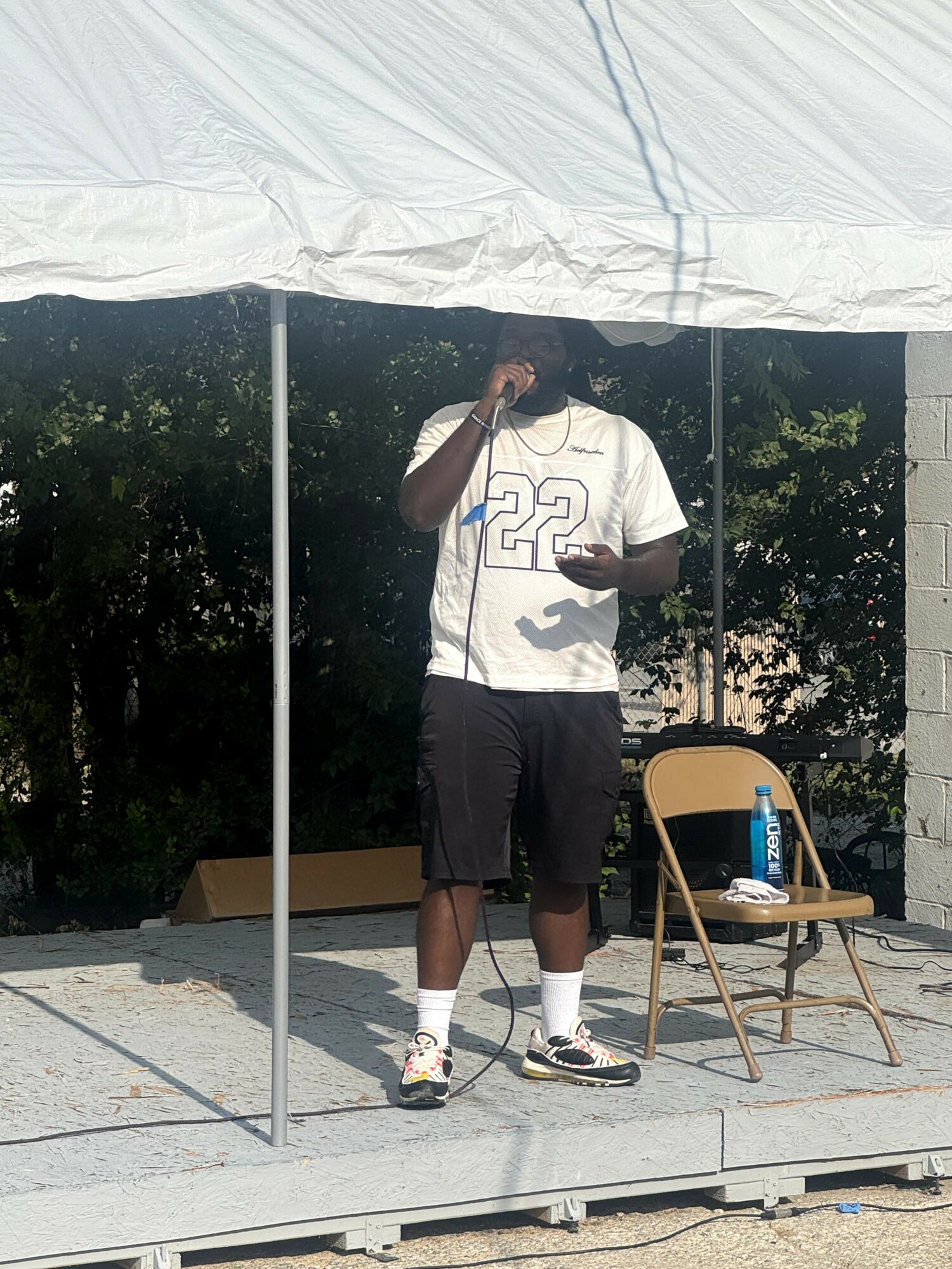Person standing on stage under a canopy, holding a phone, with a chair and water bottle nearby, outdoors.