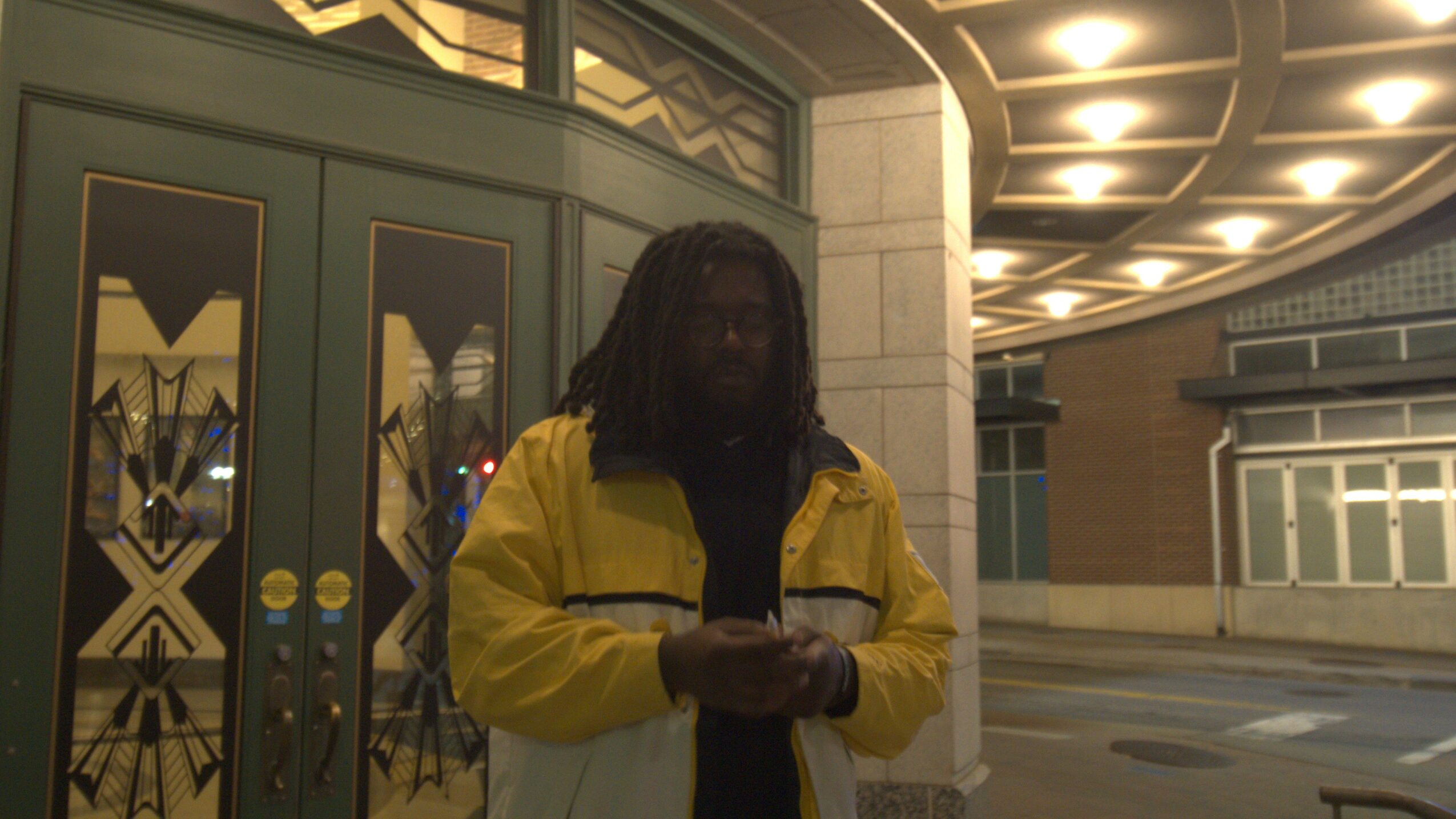 Person with dreadlocks wearing a yellow jacket standing outside near a building entrance at night.