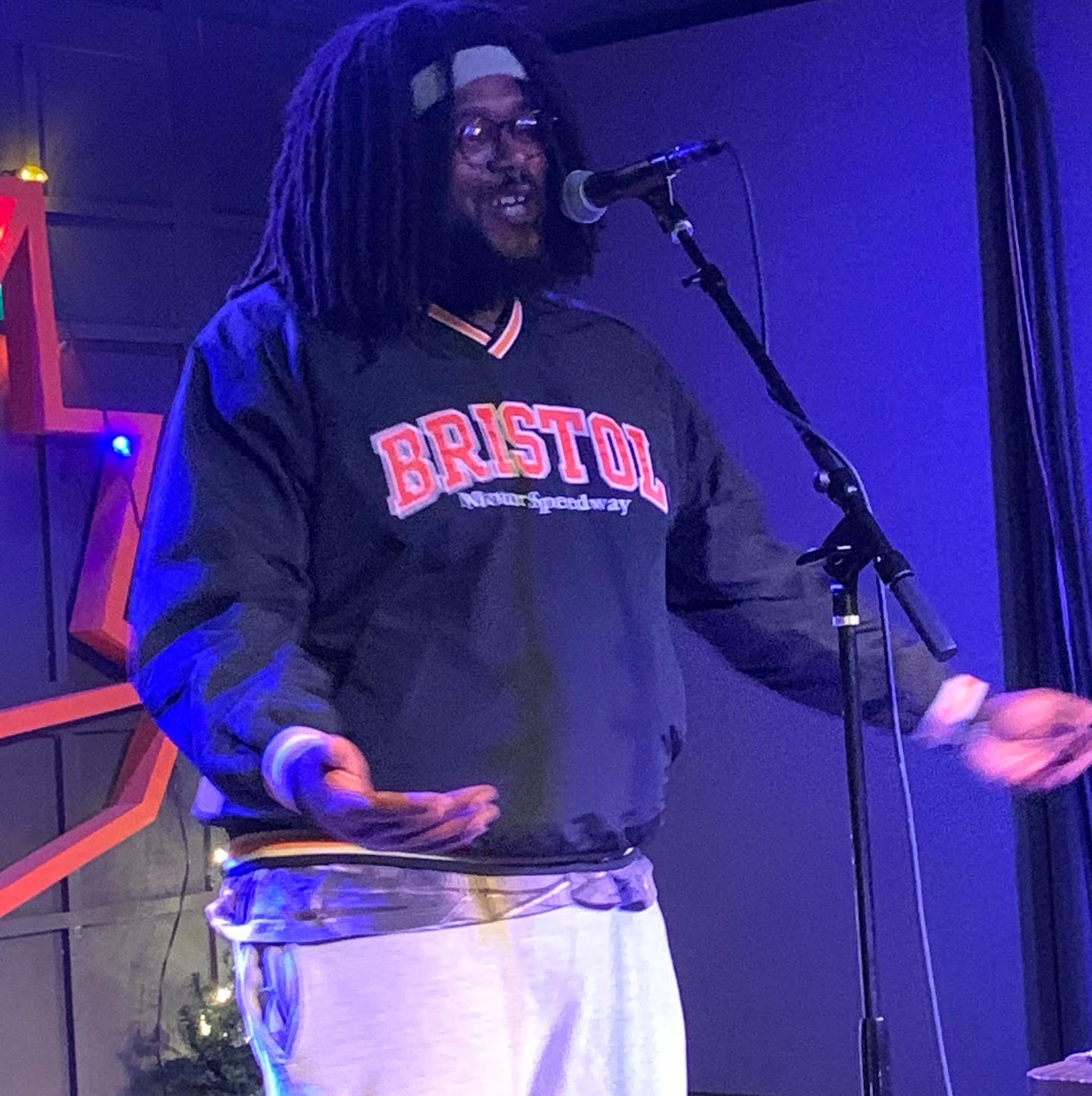 Person with dreadlocks and glasses speaking into a microphone on stage, wearing a Bristol sweatshirt and light-colored pants.