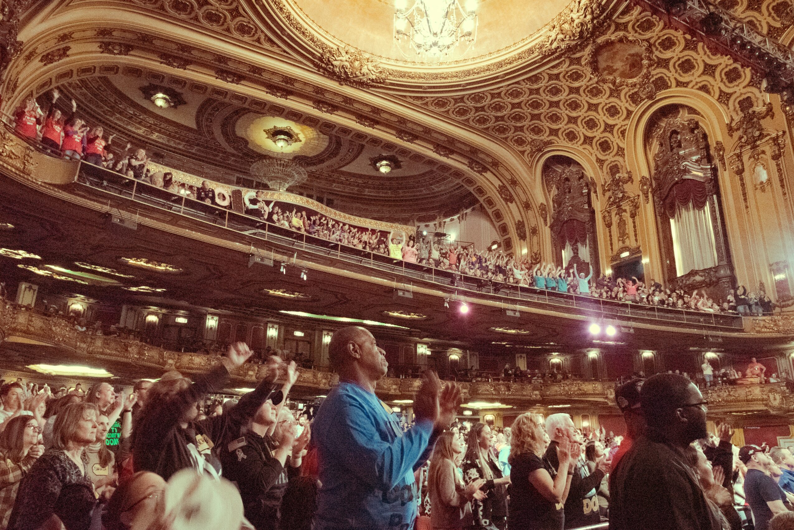 Audience seated in a grand theater with ornate architecture and a decorated ceiling, watching a performance.