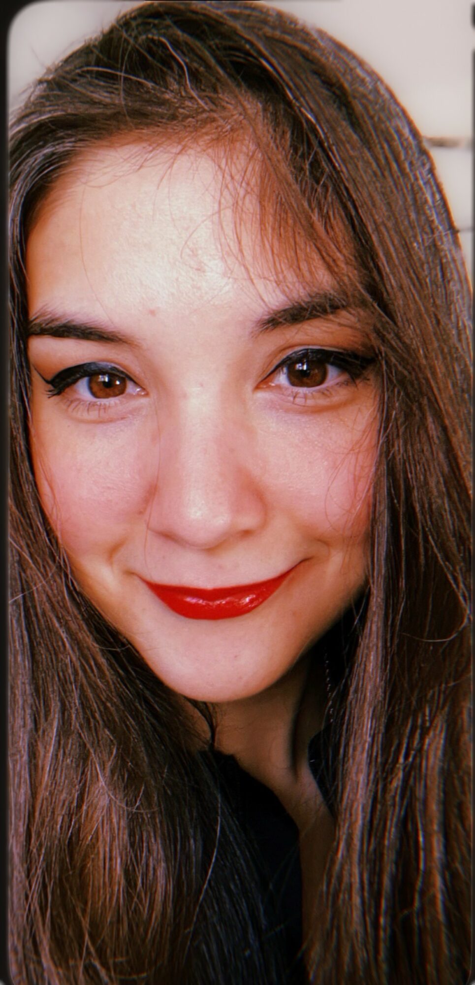 Close-up of a woman with long brown hair, smiling, wearing red lipstick, and looking at the camera.