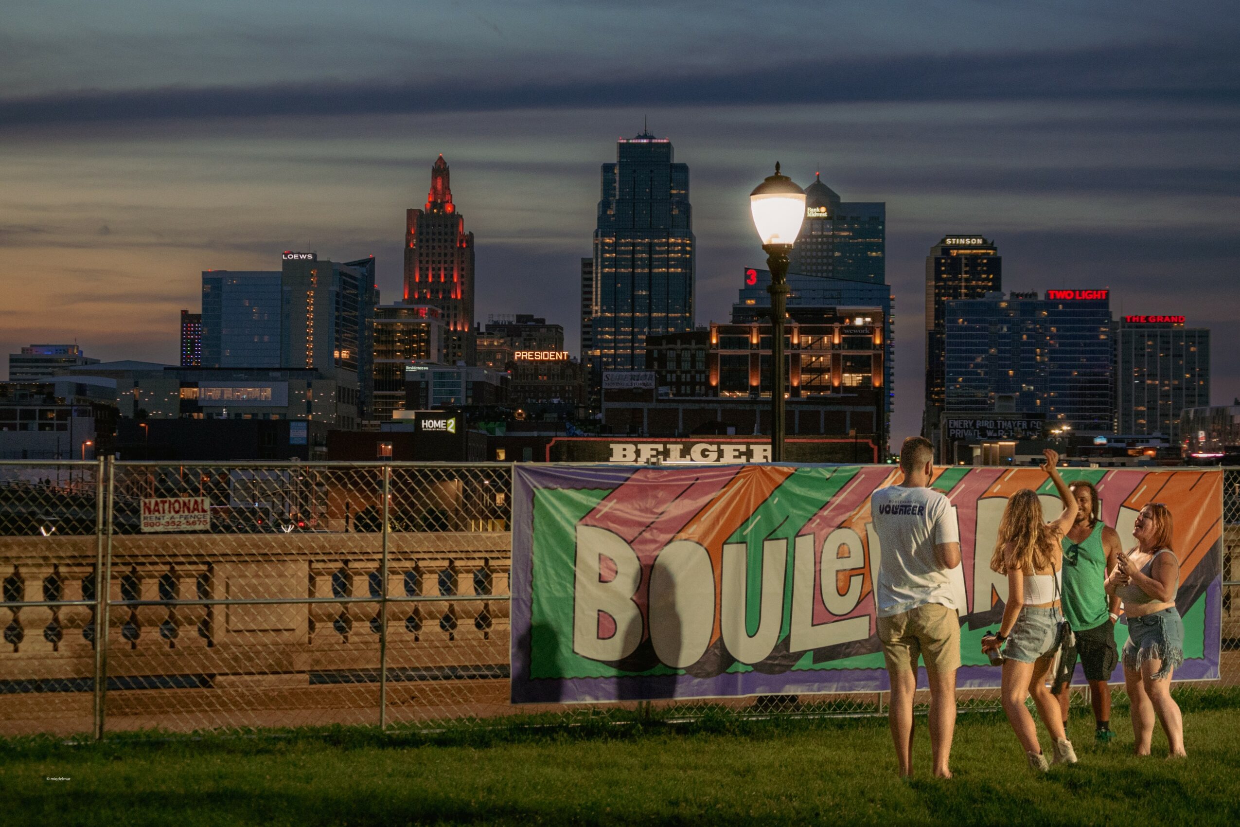 Group of people walking past a colorful banner on a bridge with a city skyline at dusk in the background.