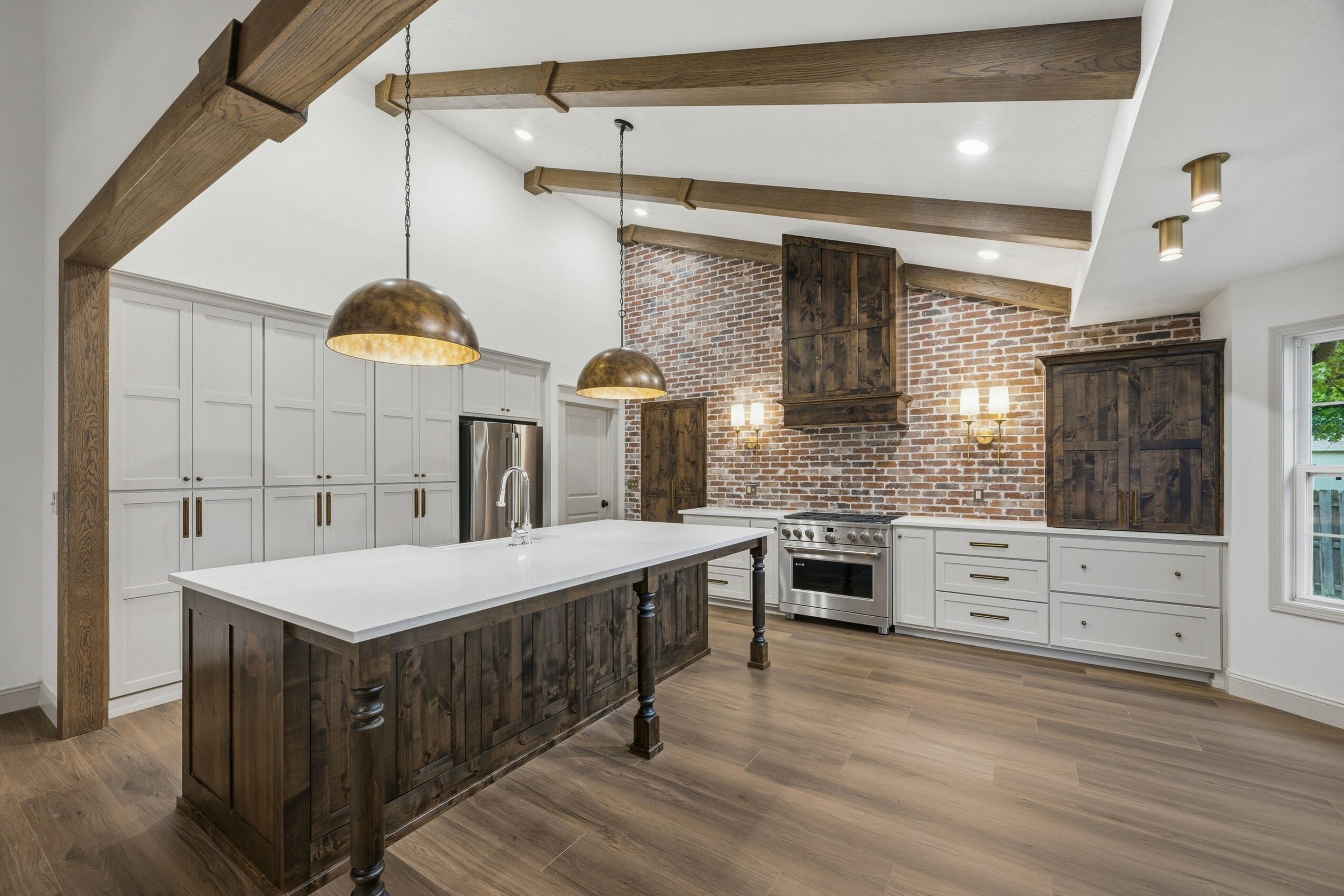Modern kitchen with white cabinets, a large island, brick wall, stainless steel appliances, and wooden beams, illuminated by pendant lights.