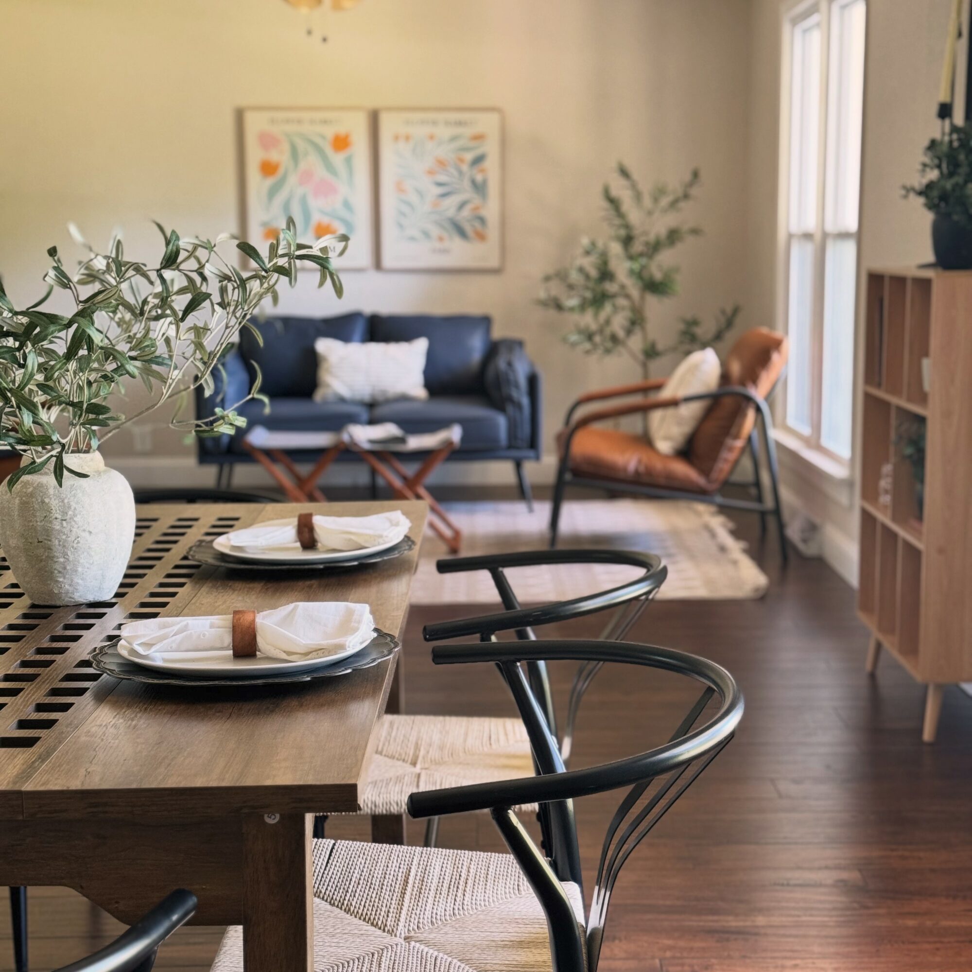 Dining table with plates and napkins, living room with sofa, armchair, and artwork, sunlight through windows, wooden floor.