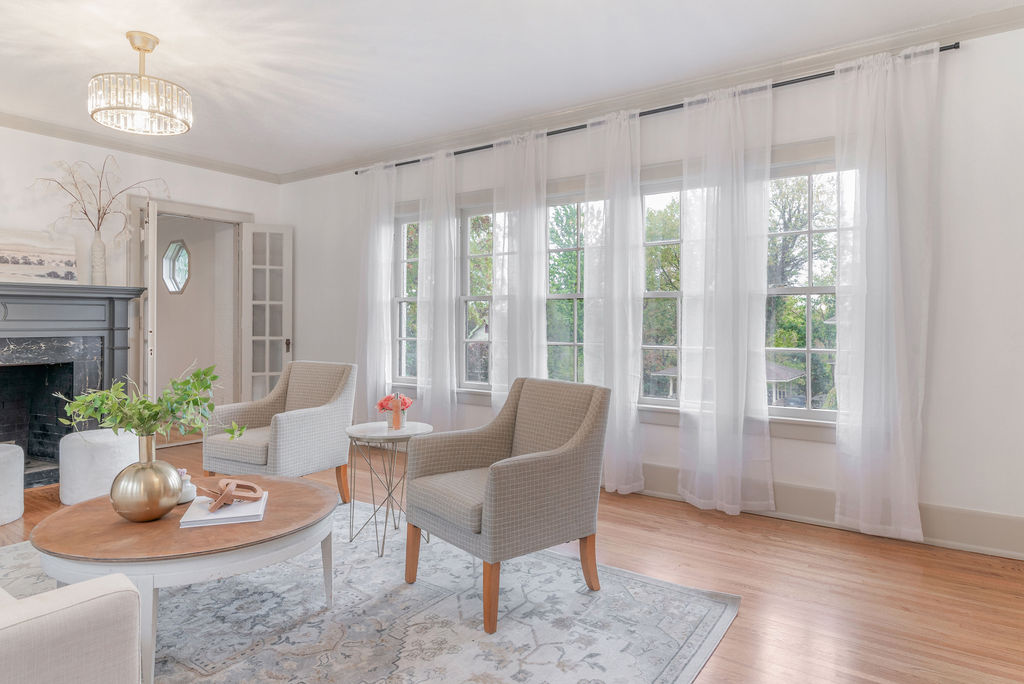 Bright living room with large windows, white curtains, beige and white chairs, fireplace, and wooden floor.