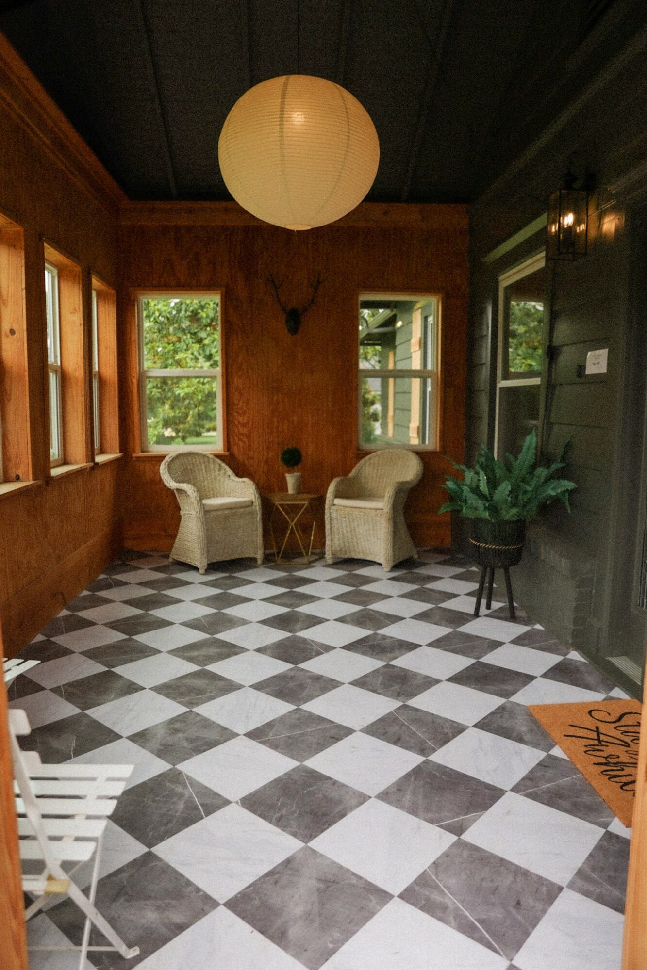 Room with checkered black and white floor, two white chairs near windows, large paper lantern ceiling light, and a potted plant.