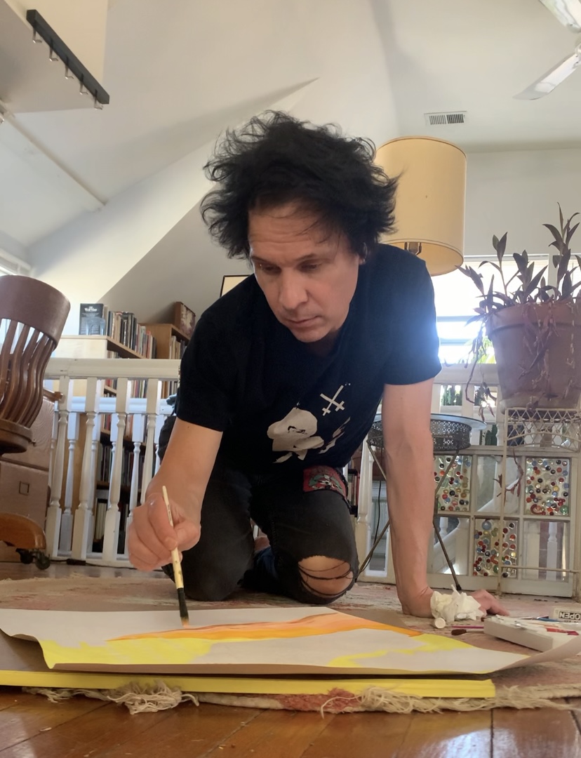 Person with curly hair kneeling on the floor, writing on large paper with a pen, in a room with books and plants.