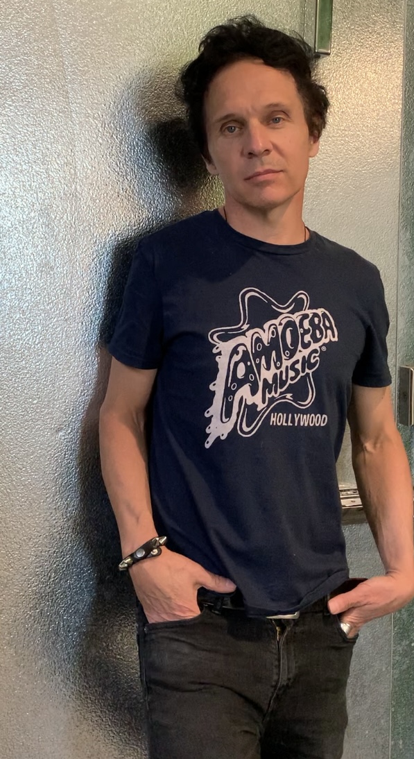 Man with dark curly hair wearing a navy T-shirt with white graphic text, standing against a textured wall.