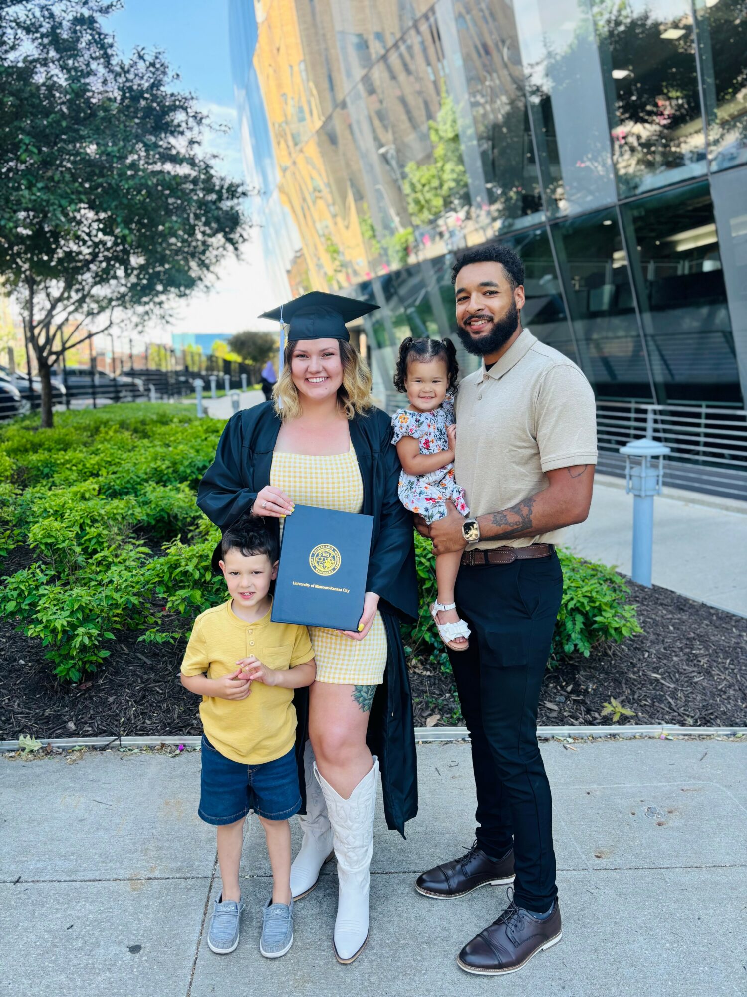 A woman in a graduation gown and cap holding a diploma, with a man and two children outdoors near a building and greenery.