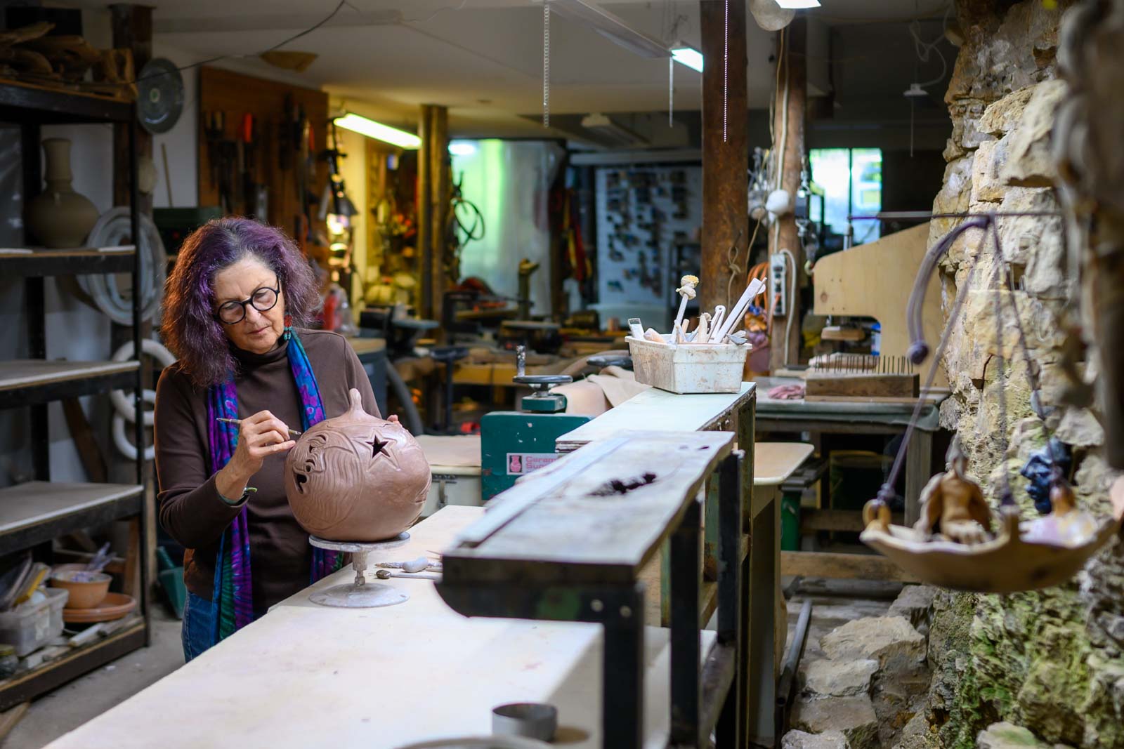 Woman with glasses and curly hair working on a clay sculpture in a workshop, surrounded by tools and shelves.