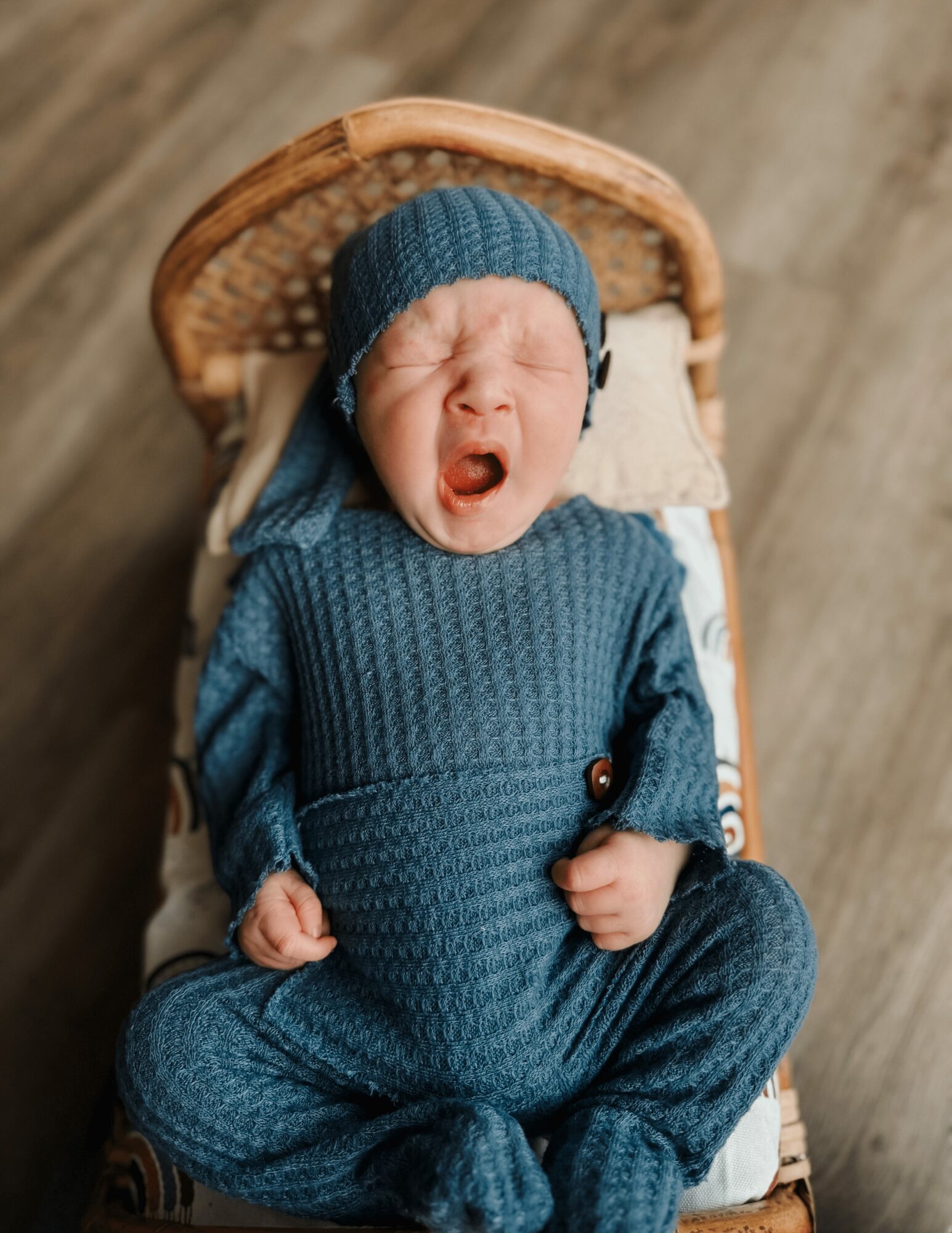 Baby in blue knit outfit and hat yawning, lying in a wicker basket on a wooden floor.