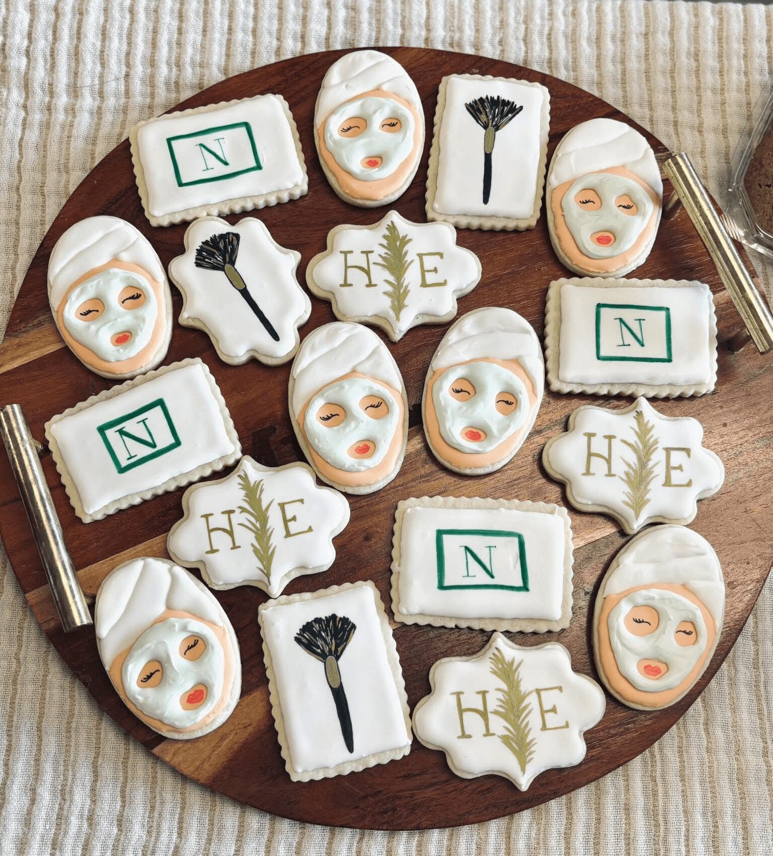 Assorted cookies with face masks, a black fan, and initials on a round wooden tray, on a striped fabric surface.