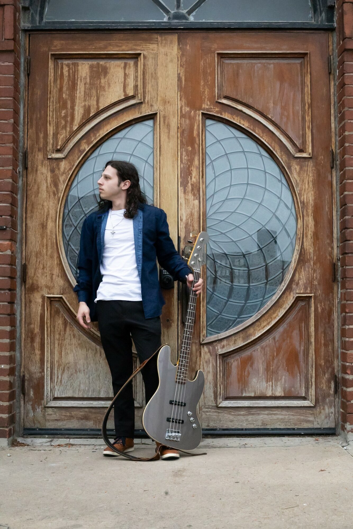 Young woman with long dark hair in a blue jacket and white shirt stands in front of a wooden door with circular glass windows, holding a bass guitar.