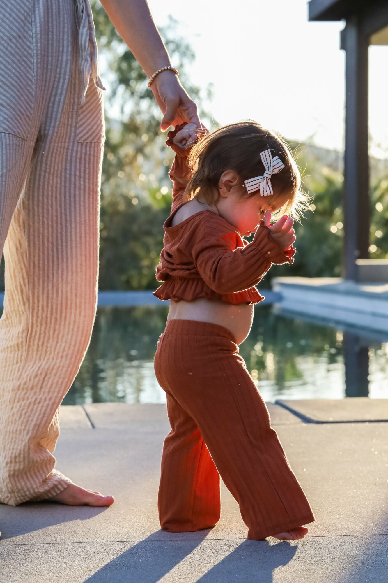 Child standing near water, holding hands with an adult, wearing brown pants and a red top, with a bow in her hair.