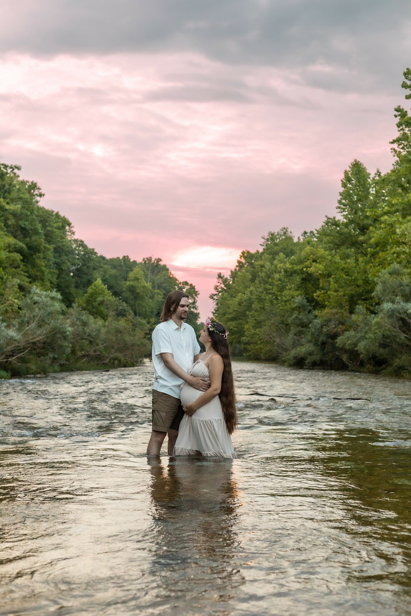 A couple stands in a river, embracing, surrounded by trees with a pink and purple sky above.