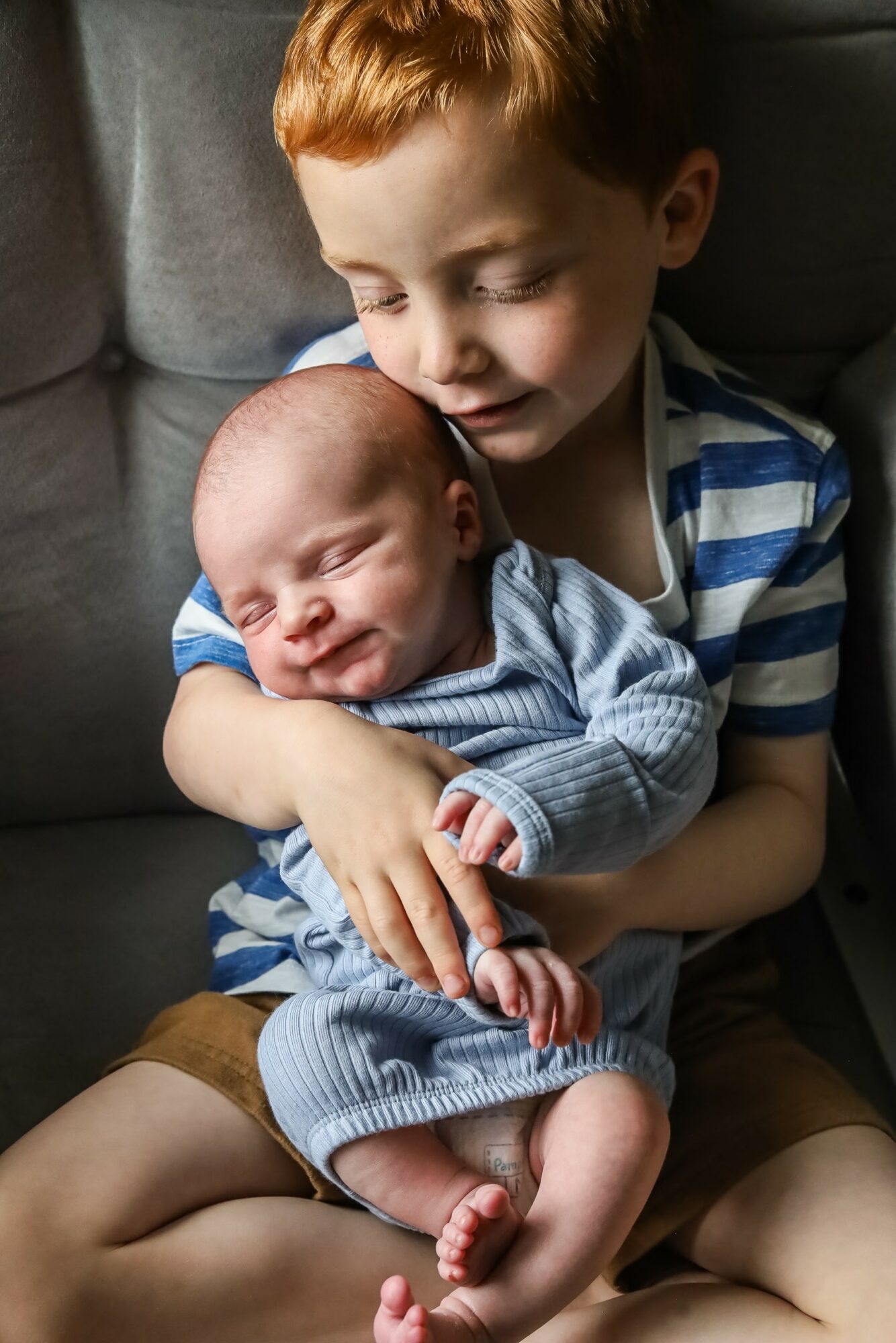 A young boy holding a newborn baby, both sitting on a gray couch, with the boy smiling gently.