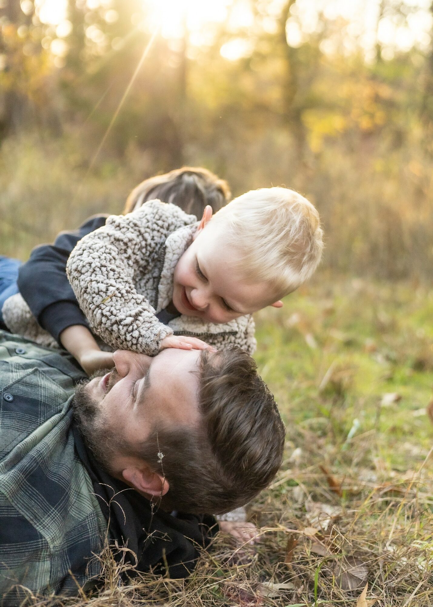 Child and adult lying on grass outdoors, smiling and playing together in sunlight, surrounded by trees in autumn.
