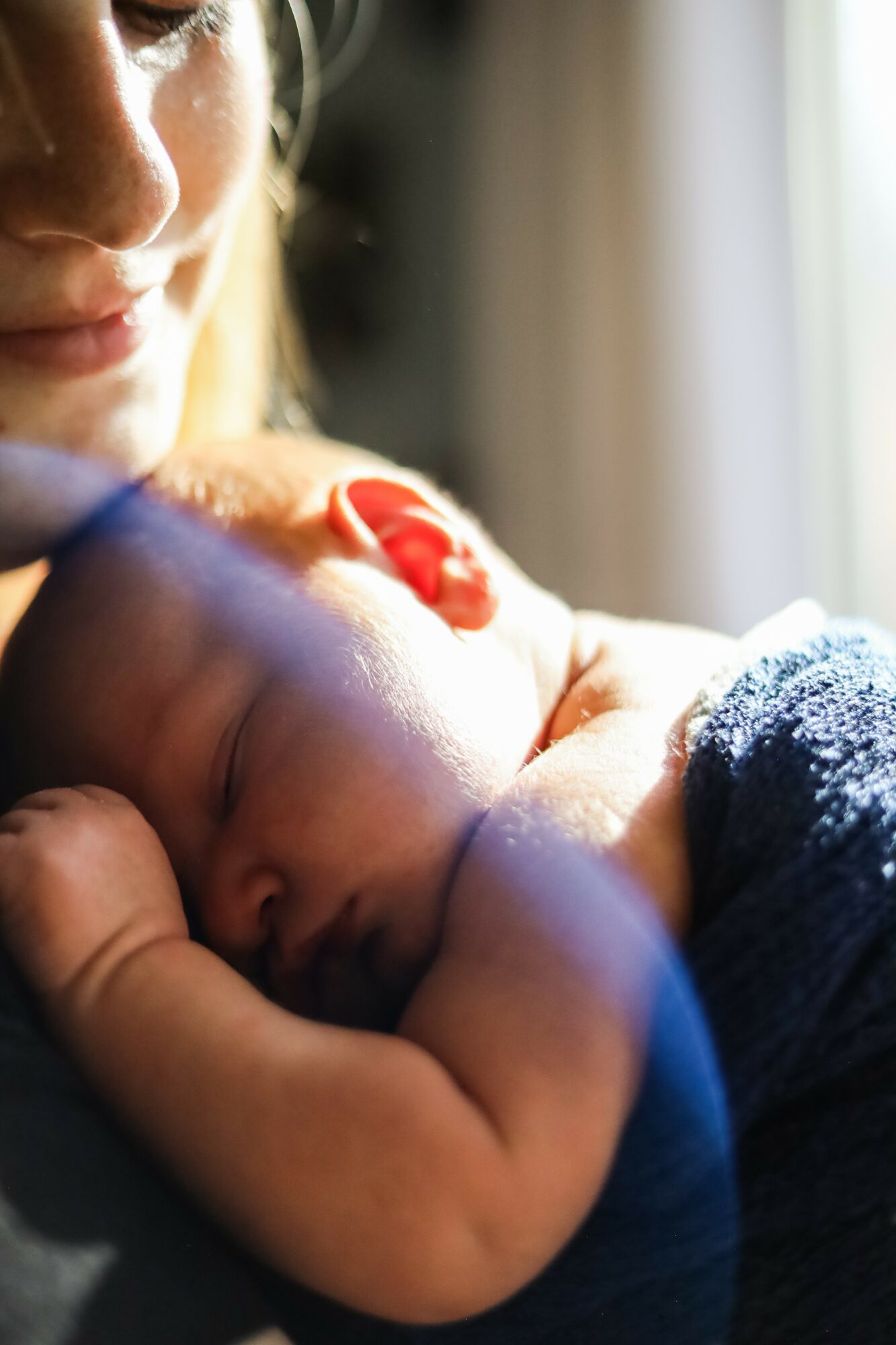 Person holding a sleeping baby with a red clip on baby's head, near a window, soft lighting.