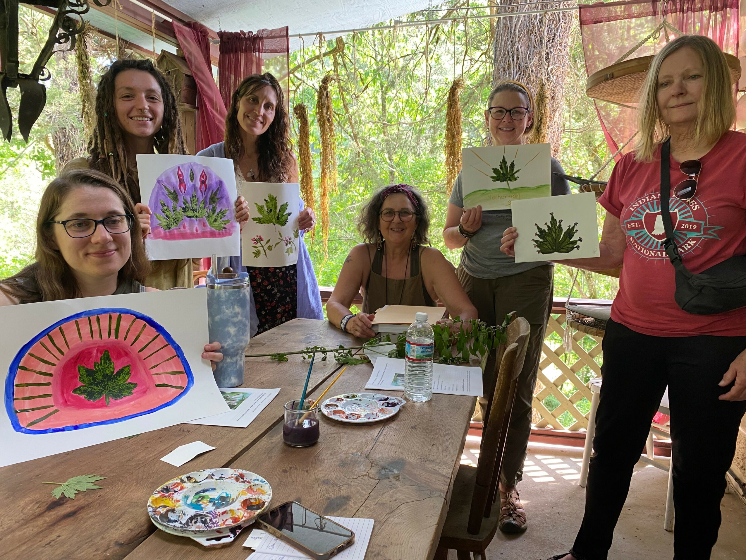 Six women display artwork with plant images in a bright, outdoor setting with trees and pink curtains, sitting and standing around a table.