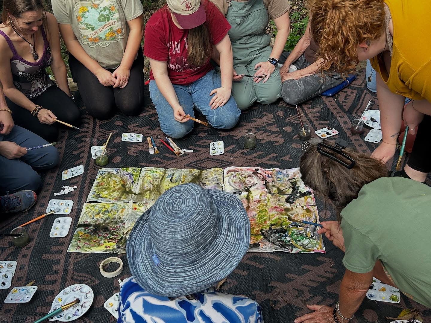 Group of people sitting on a carpet, working on a large, colorful painting with art supplies around them.