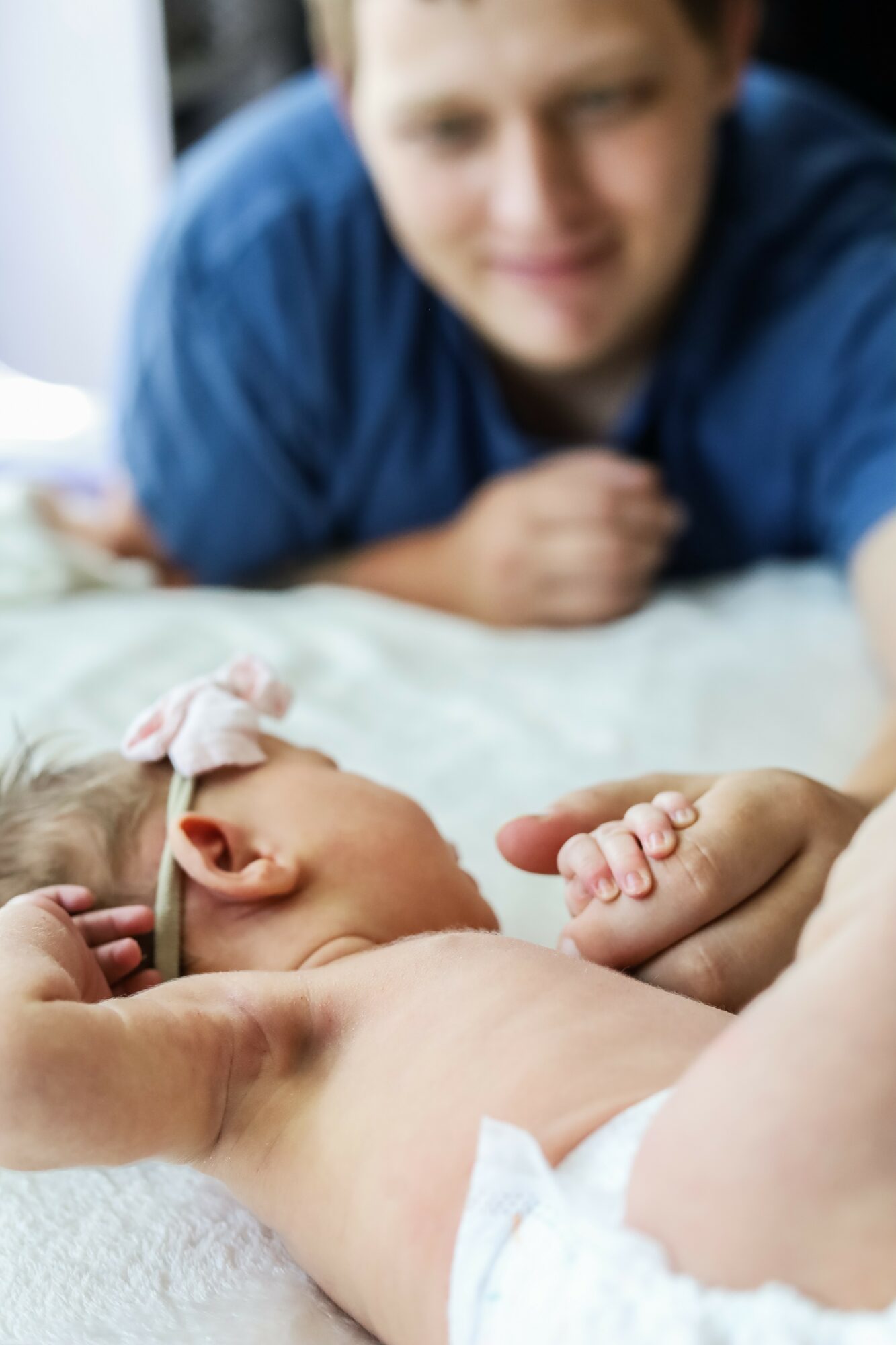 A person with a blue shirt looks at a newborn baby lying on a bed, holding the baby's hand.