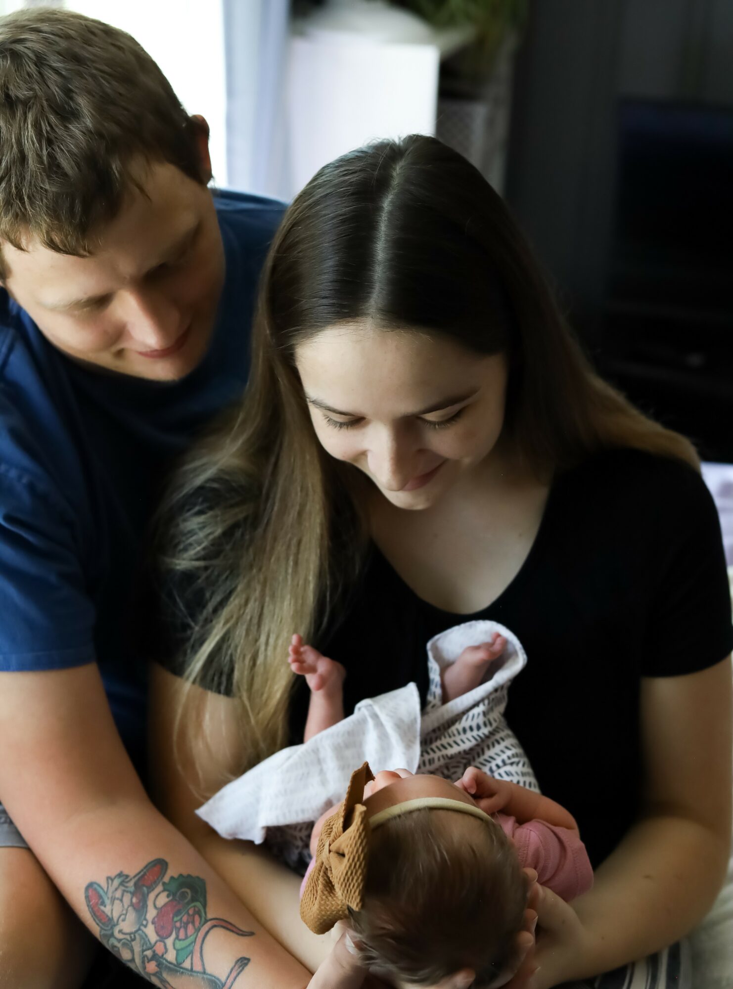 Two people holding a newborn baby indoors, smiling and looking at the baby, with a dark background and a window nearby.